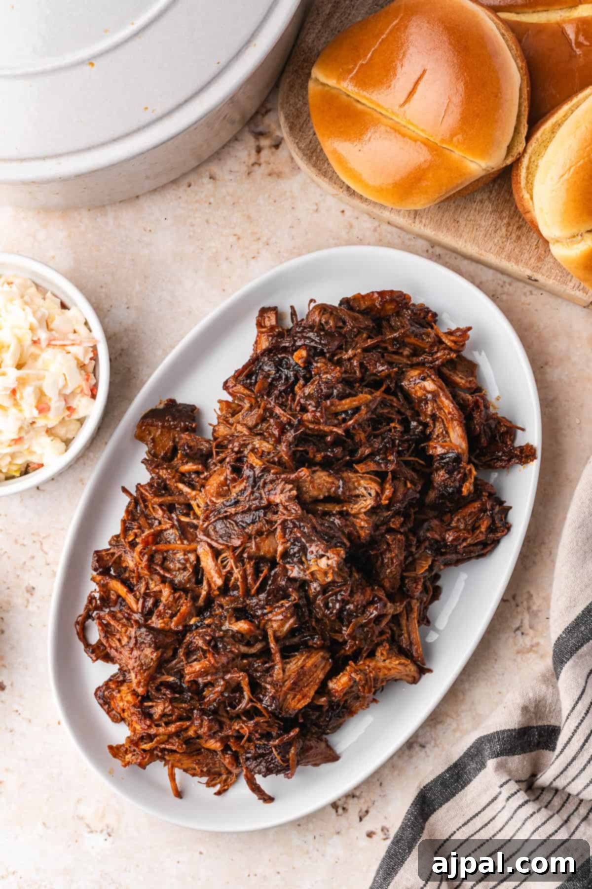 A delectable display featuring a Dutch oven's masterpiece: an oval plate of pulled pork complemented by a bowl of tangy coleslaw, round bread buns, and a charming striped cloth on a light textured surface.