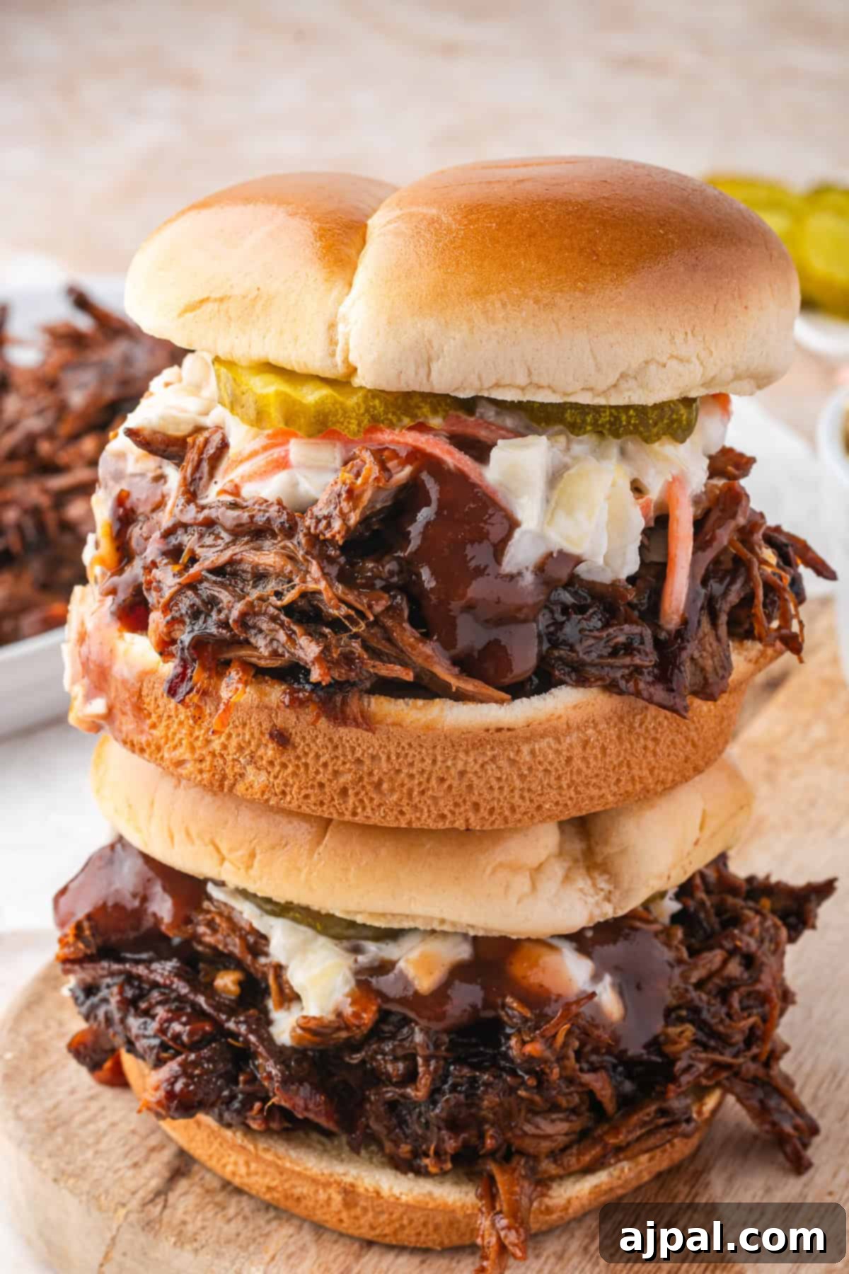 Two stacked BBQ pulled beef sandwiches, rich with flavor and topped with creamy coleslaw and tangy pickles, rest enticingly on a wooden board. These mouthwatering delights rival even the tastiest Dutch Oven Pulled Pork recipes.