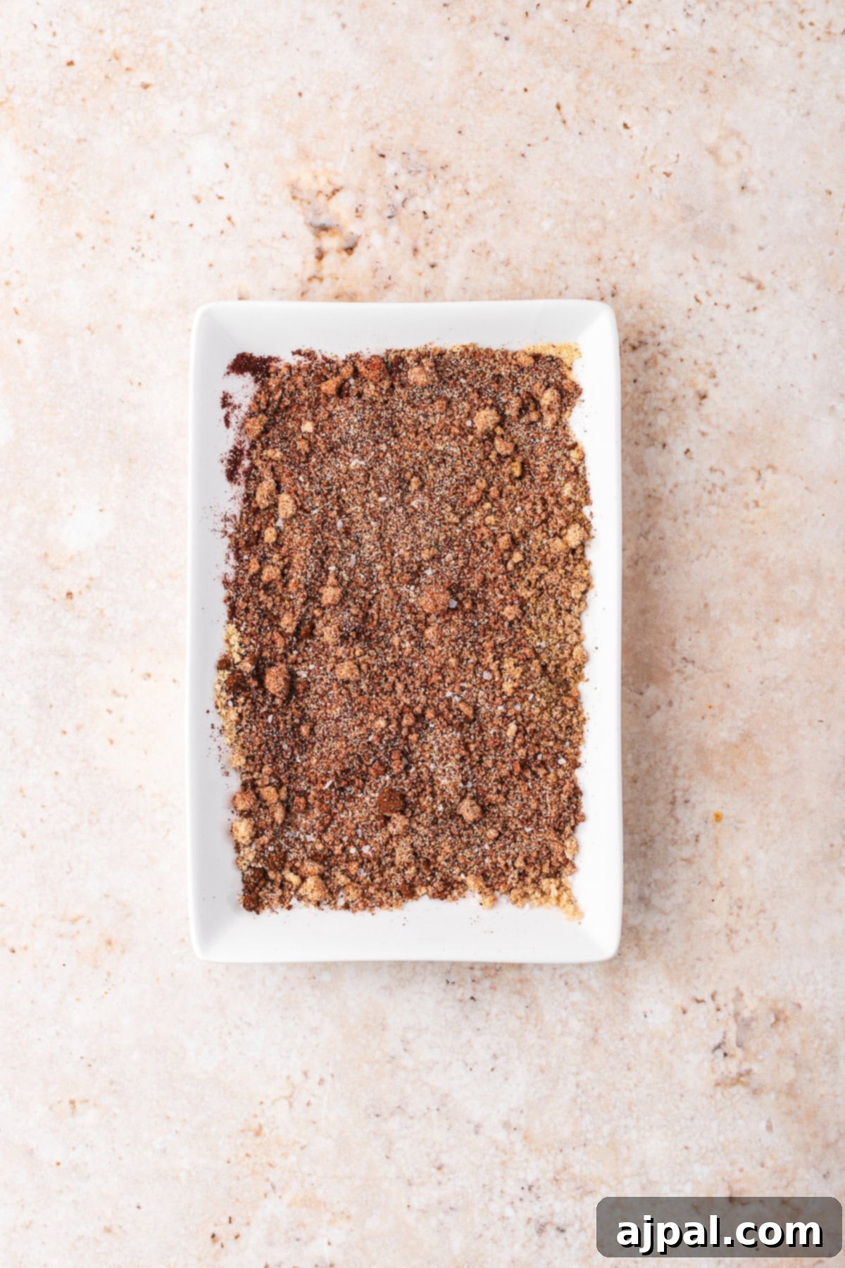 A rectangular white dish filled with ground spices sits on a speckled beige surface, perfect for seasoning your favorite pulled pork recipe in a Dutch oven.