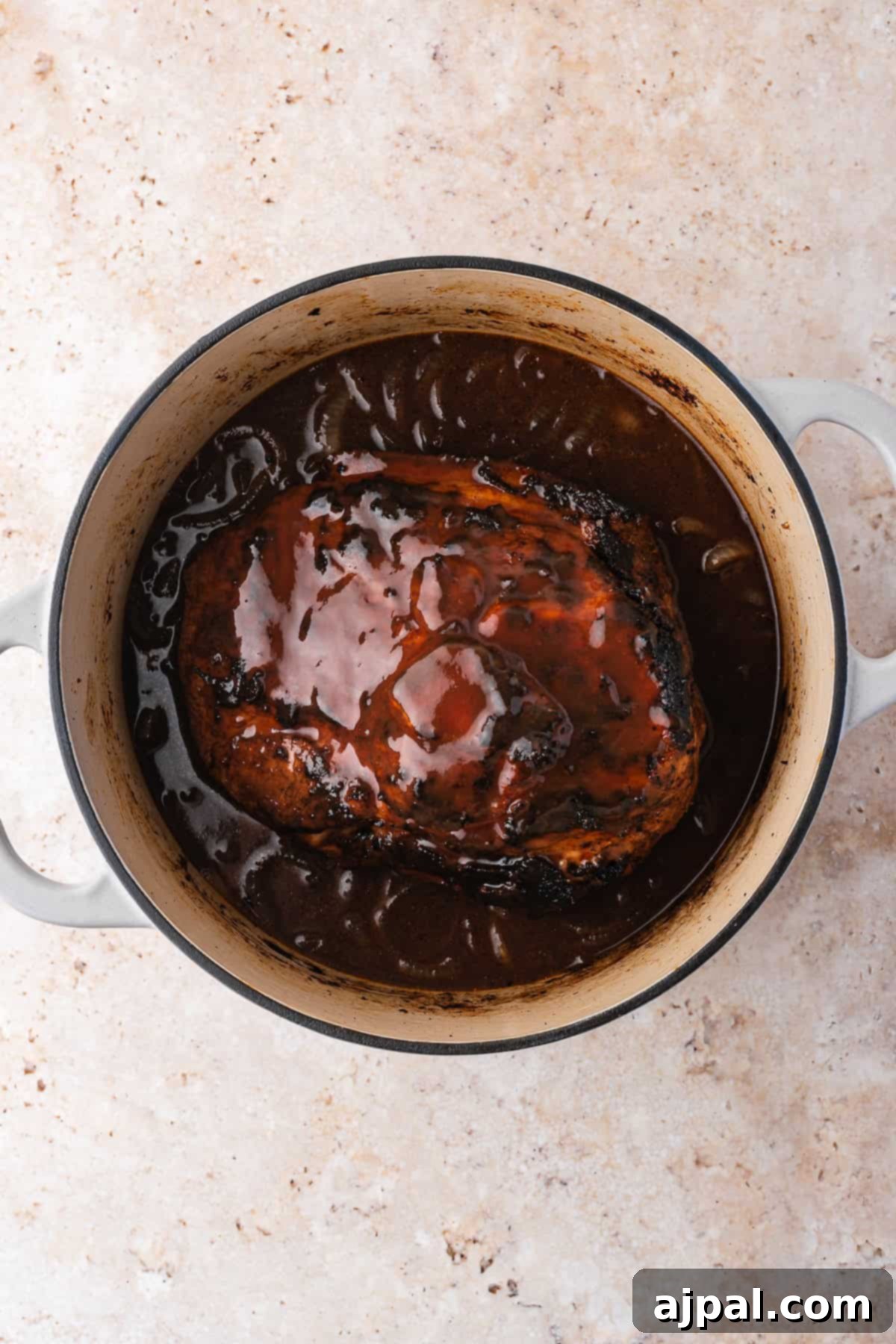 A succulent piece of meat, resembling pulled pork, is bathed in a rich brown sauce inside a round Dutch oven, resting on a light-colored textured surface.