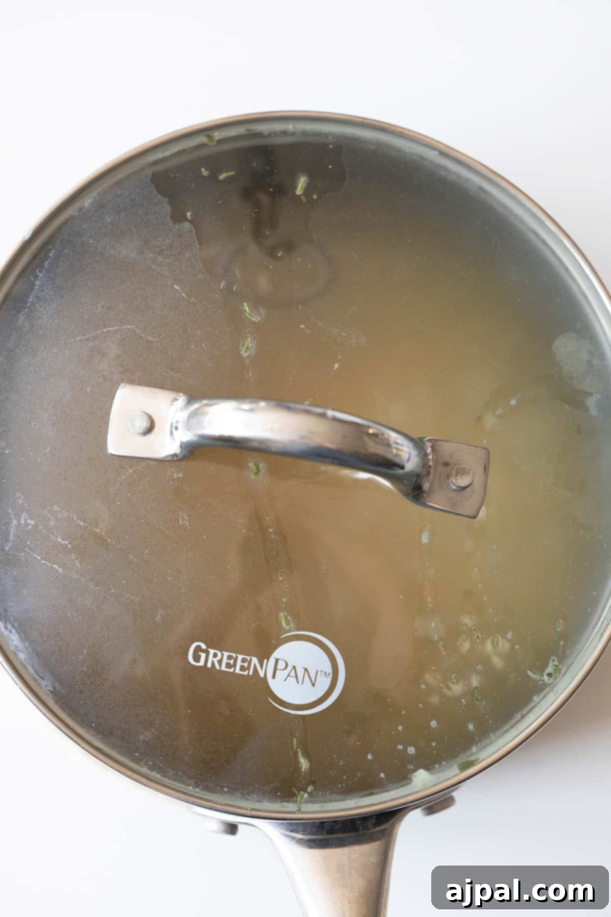 A stainless steel pot with a glass lid on, filled with simmering clear liquid—a perfect start for a couscous recipe. The handle is metal, and the lid has "GreenPan" printed on it, partially fogged by the gentle heat of cooking.
