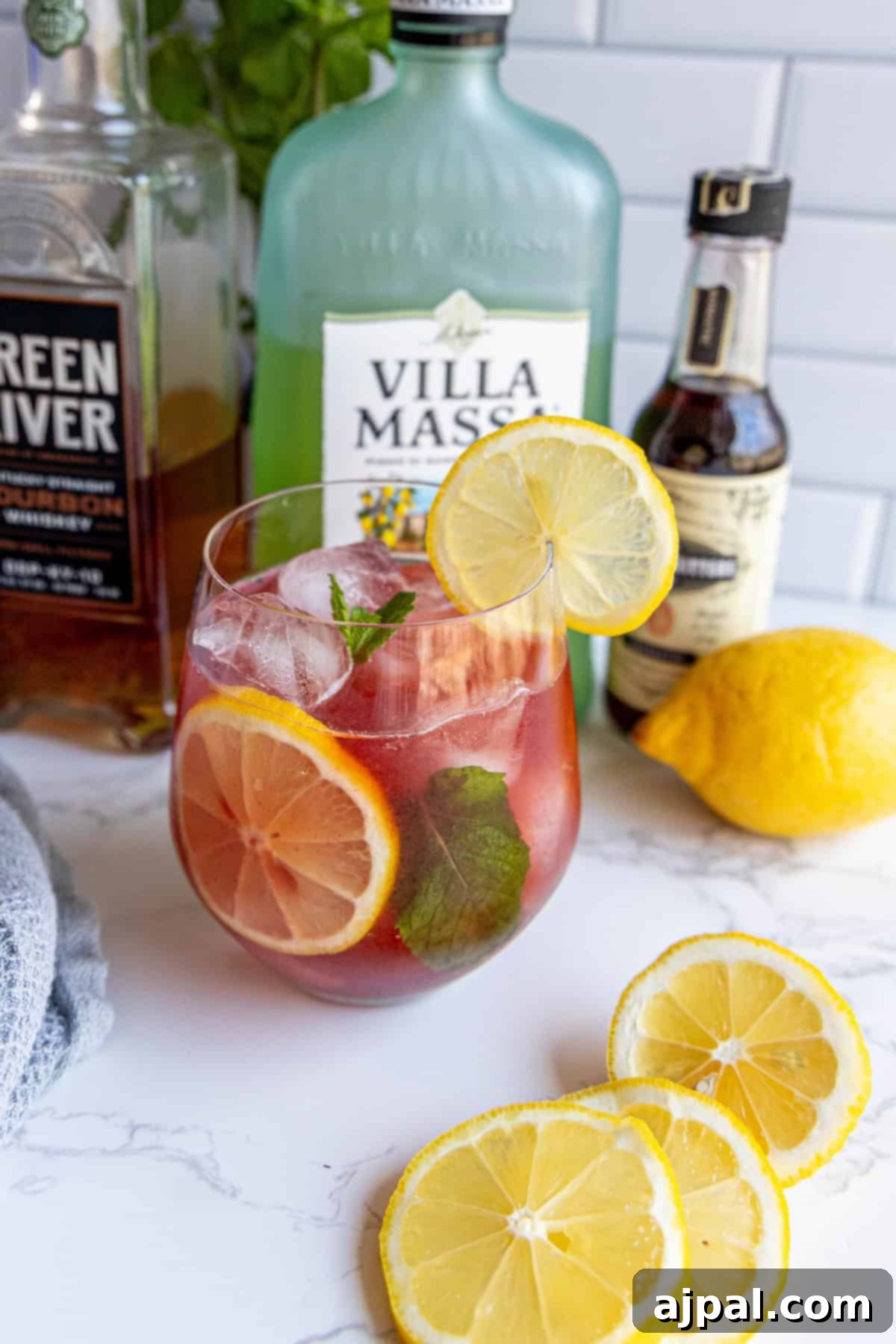 A glass of iced cocktail garnished with lemon slices and mint, surrounded by bottles and a whole lemon on a marble surface.