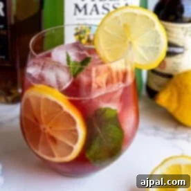 A glass of iced red cocktail garnished with lemon slices and mint leaves. Bottles and a whole lemon are visible in the background.