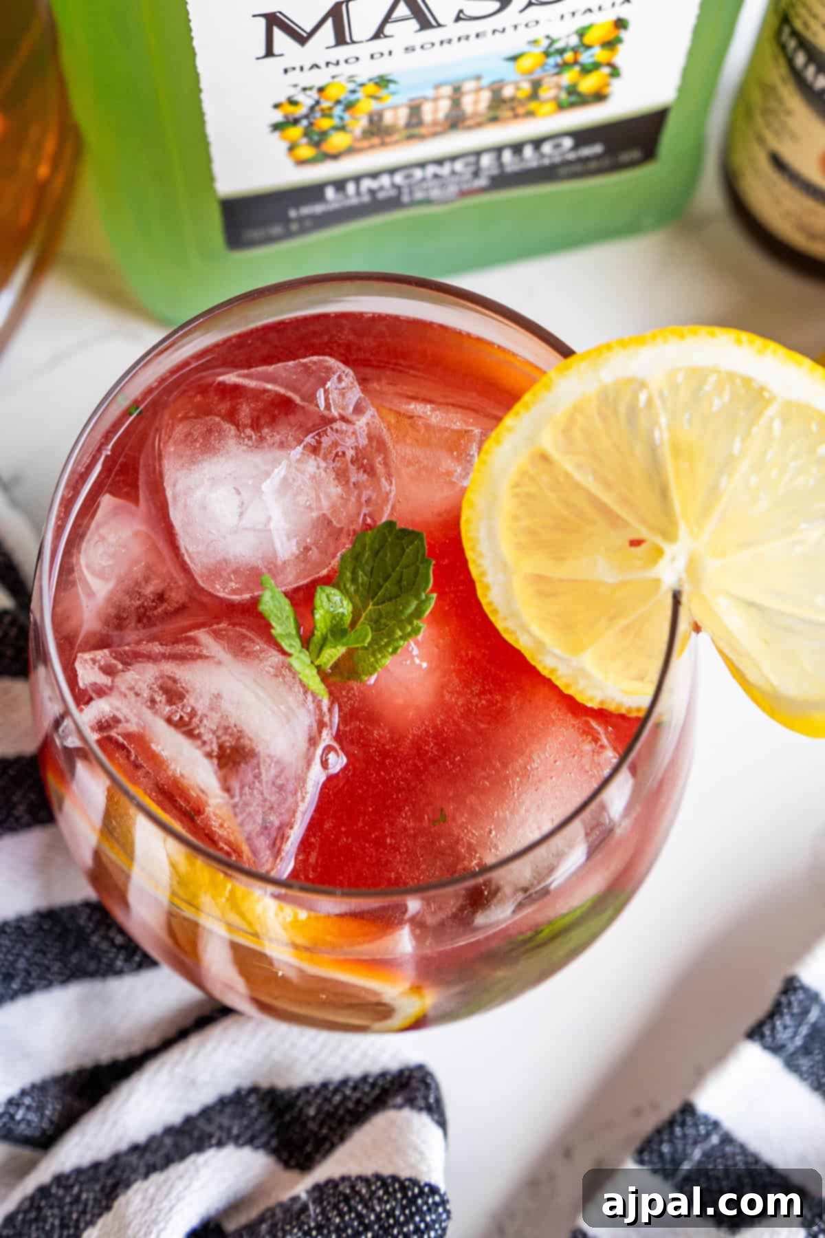 A glass of pink cocktail with ice, garnished with a lemon slice and mint leaf, next to a bottle labeled "Limoncello." A striped cloth is partially visible at the bottom.