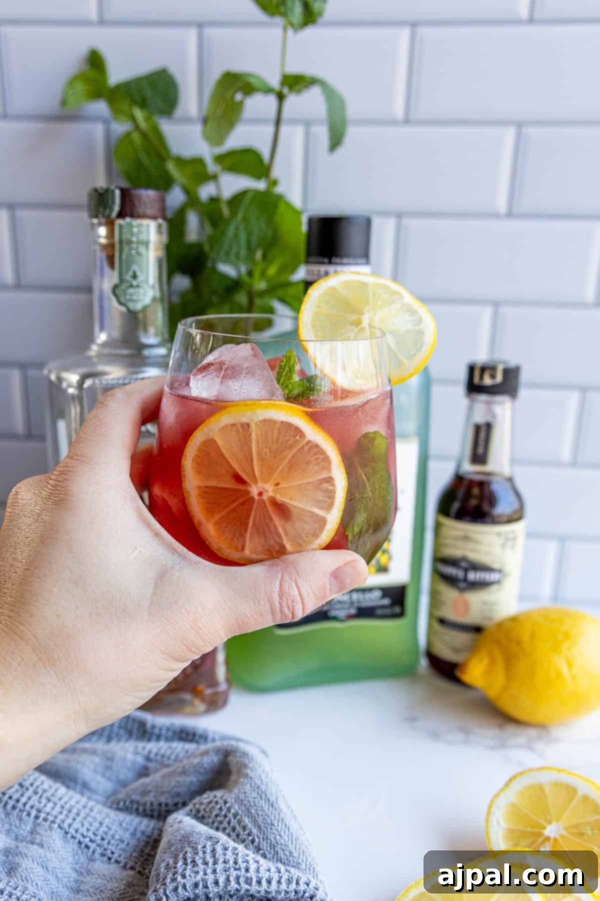 Hand holding a cocktail with a lemon slice, in front of a bottle of liquor, bitters, and a lemon on a countertop.