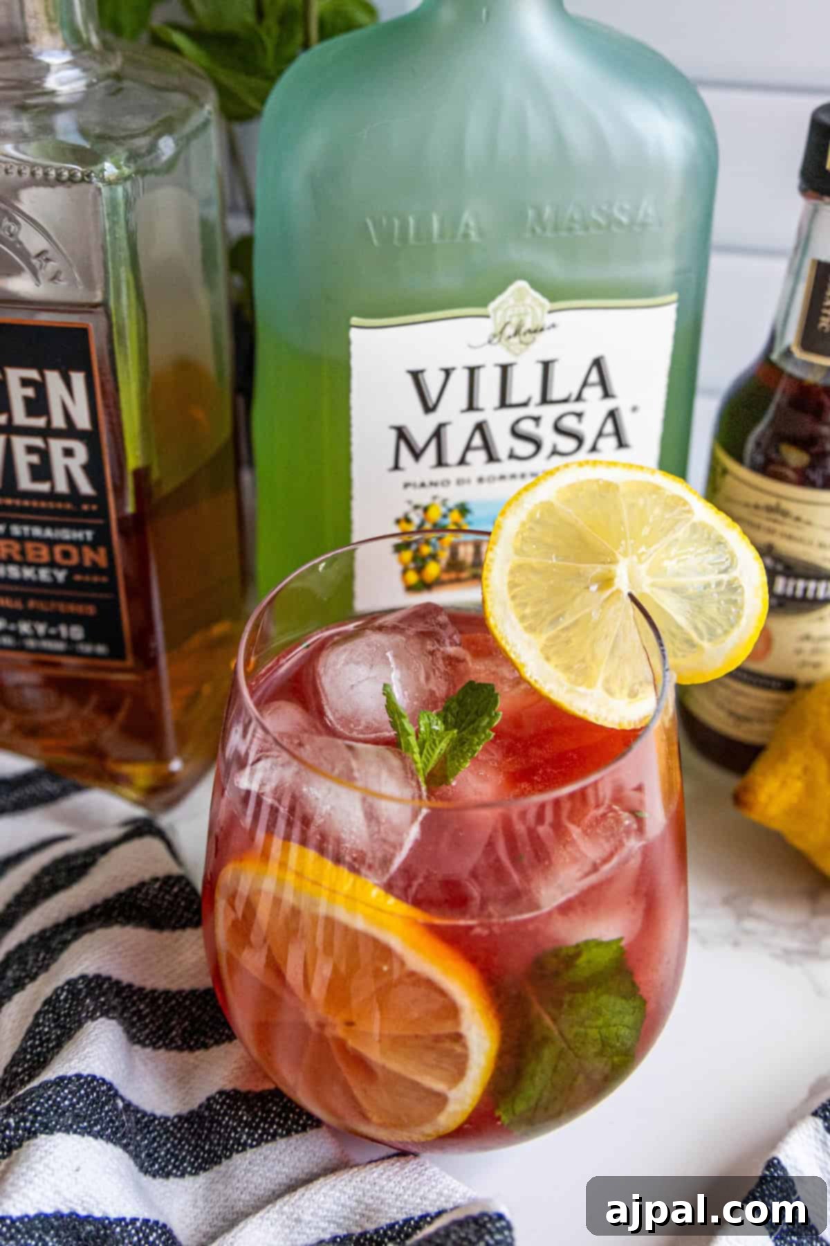 A cocktail with ice, lemon, and mint in a glass. Bottles of liqueur and bourbon are in the background, along with a striped cloth.