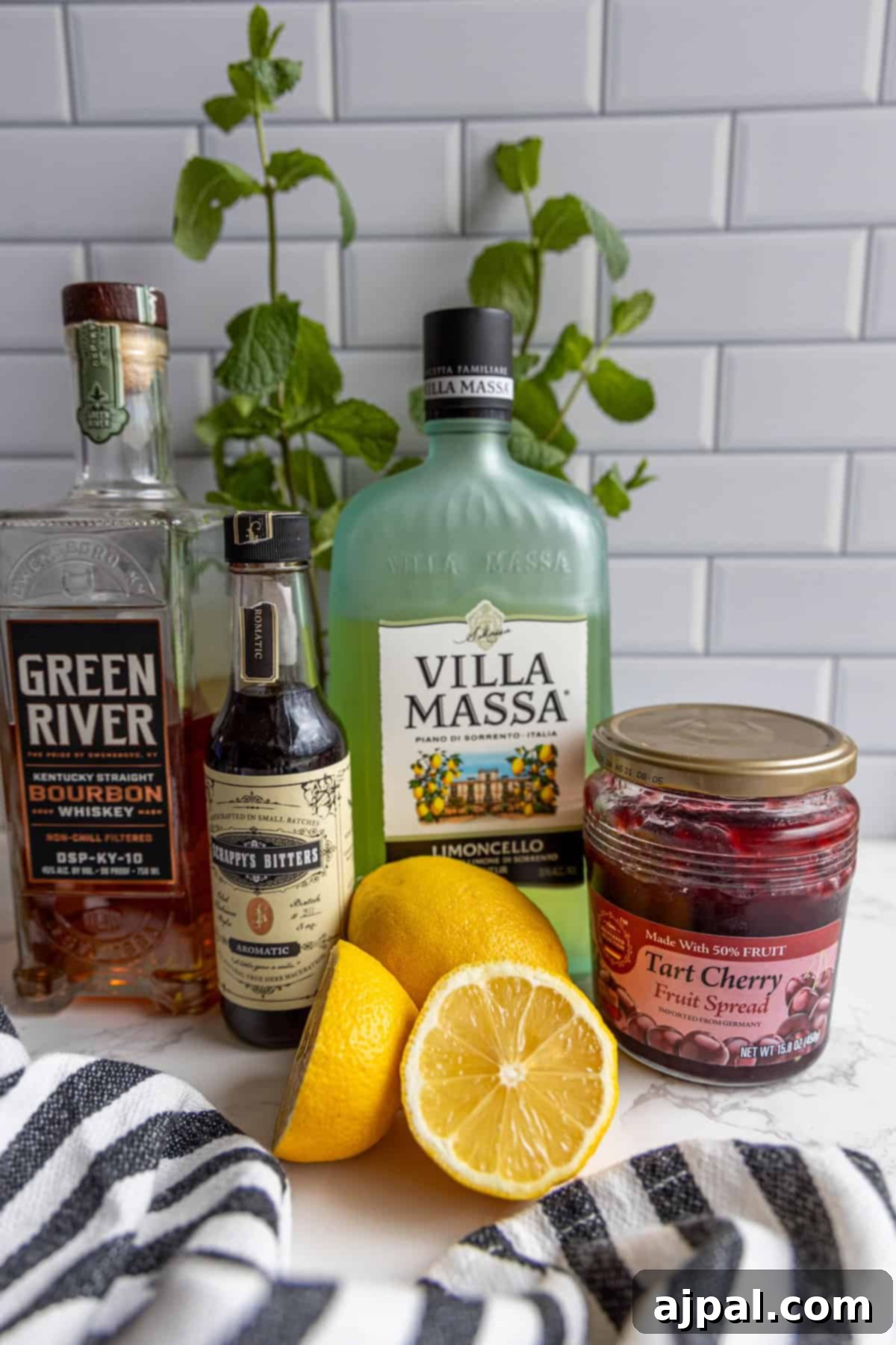Bourbon, limoncello, bitters, tart cherry spread, and a sliced lemon are on a countertop with a striped cloth and mint plant in the background.