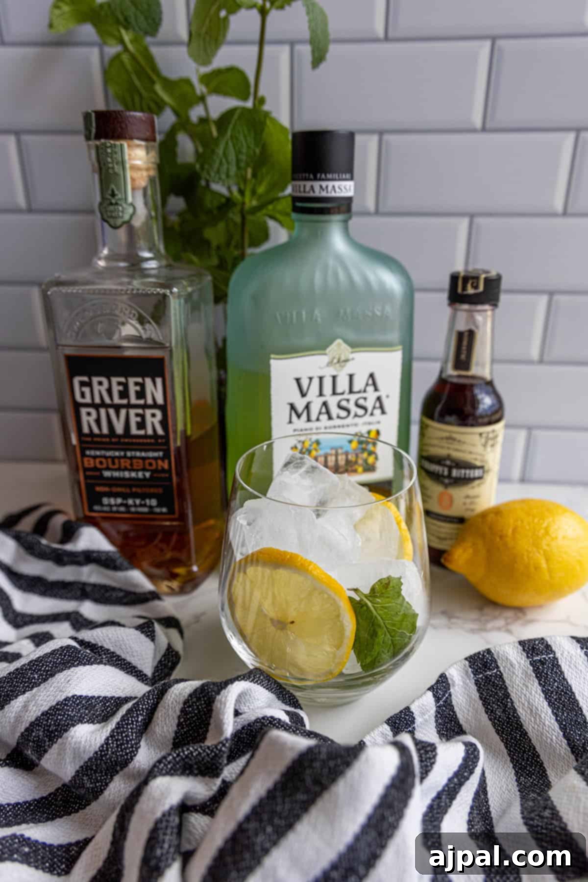 A cocktail with lemon and mint is placed in front of bottles of bourbon, limoncello, bitters, and a lemon, on a striped cloth with a tiled backsplash.
