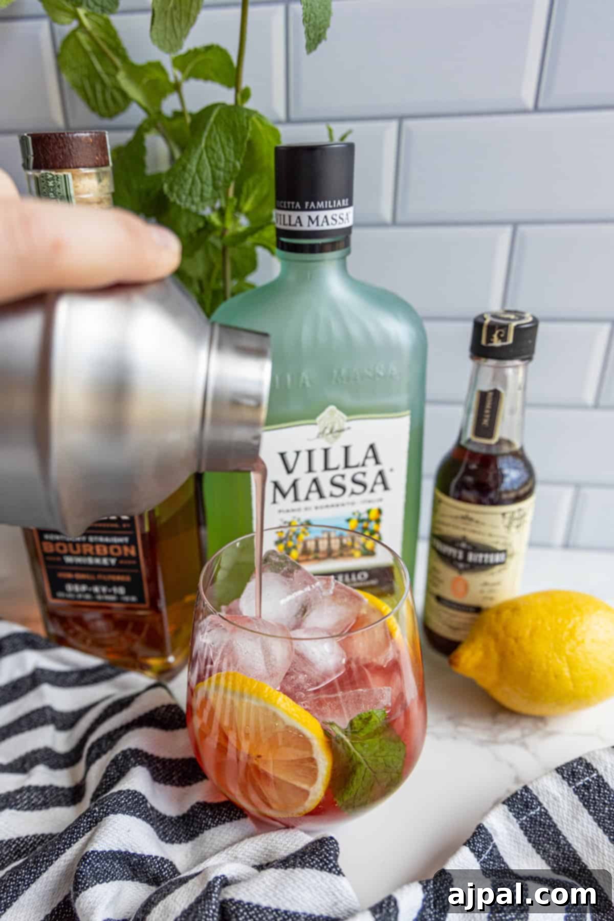 A person pours a pink cocktail into a glass with ice, orange slice, and mint. Bottles of liquor and bitters, a lemon, and a striped cloth are in the background.