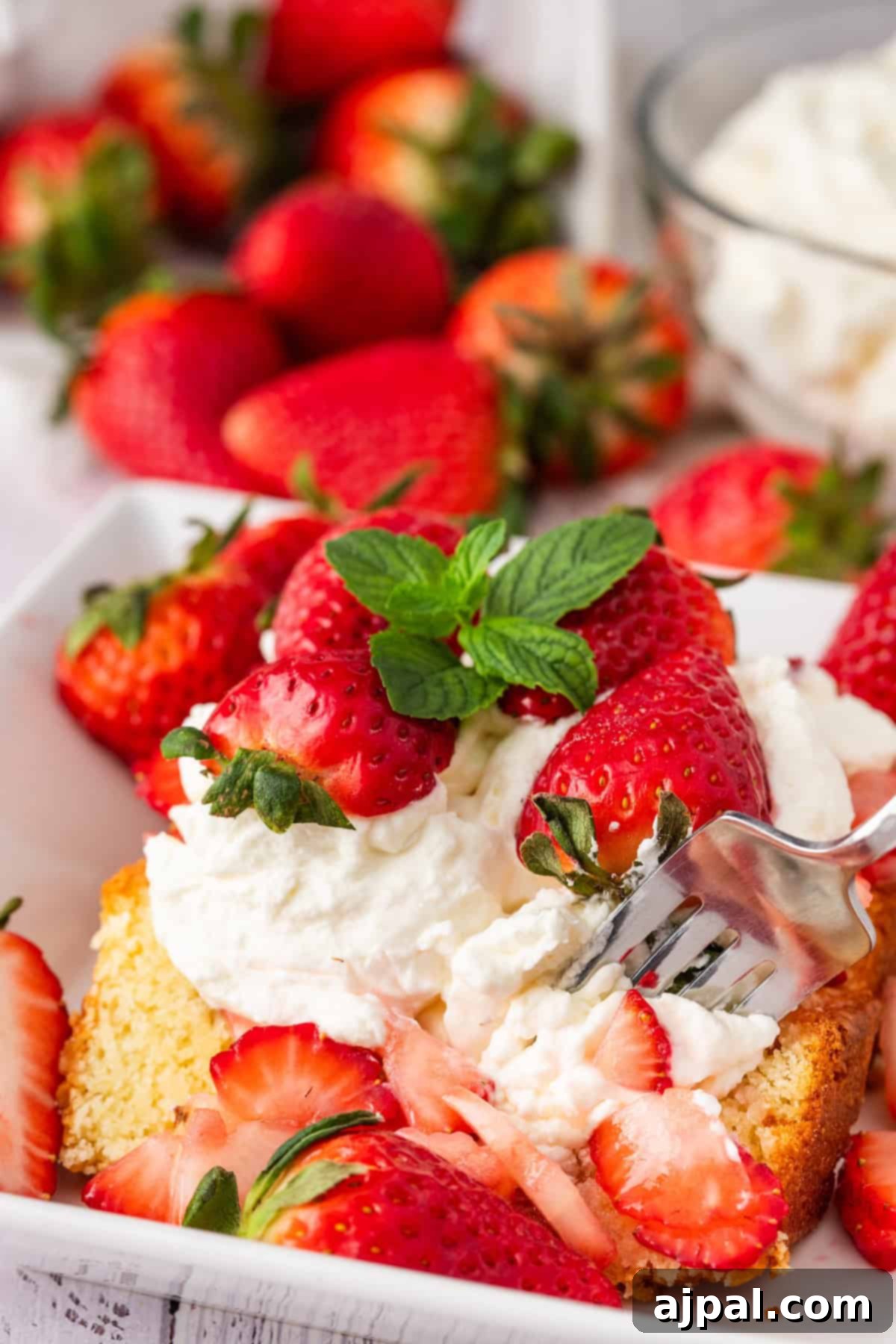 Summer Berry Shortcake 11 A hand adds whipped cream to a slice of strawberry shortcake on a square white plate, perfectly topped with fresh strawberries. A bowl of whipped cream sits beside it on the light wooden table.