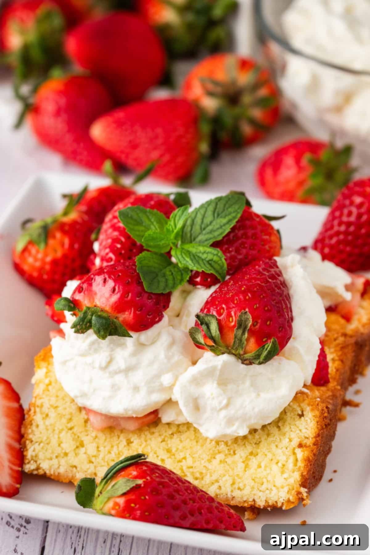 Summer Berry Shortcake 4 A slice of strawberry shortcake topped with whipped cream and fresh berries, garnished with a mint leaf on a white plate. Additional strawberries are artfully scattered around.