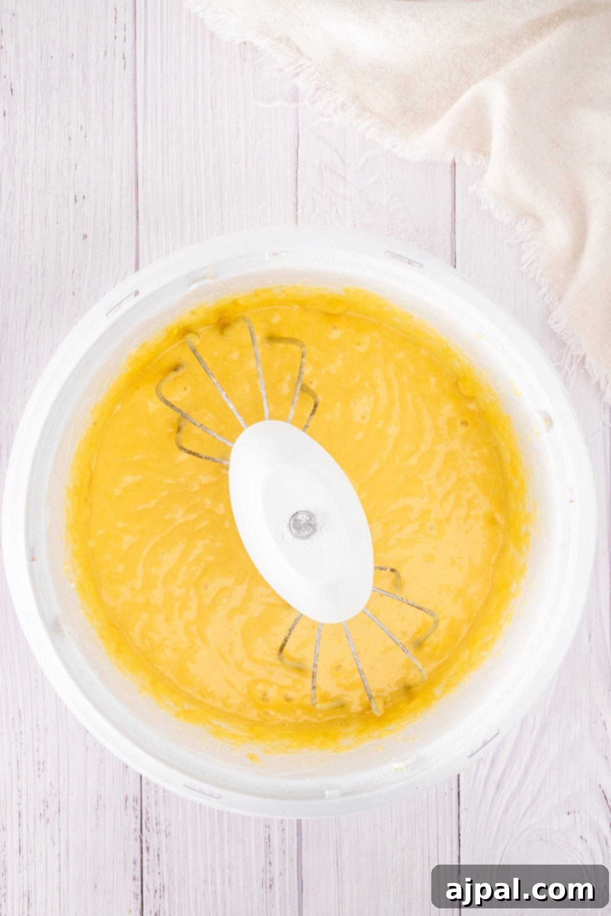 Summer Berry Shortcake 6 A bowl of yellow cake batter with a whisk attachment sits on a light wooden surface, hinting at the creation of a delightful strawberry shortcake. Nearby, a cloth rests casually, ready to assist in crafting this sweet treat.