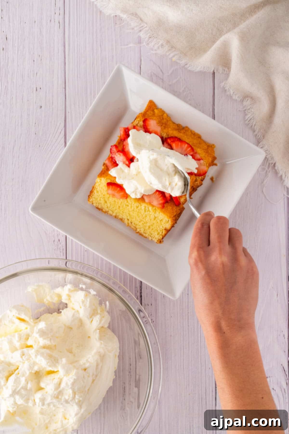 Summer Berry Shortcake 10 Top view of a white mixing bowl with creamy batter for strawberry shortcake being mixed by metallic beaters, set on a light wooden surface with a beige cloth at the top edge.