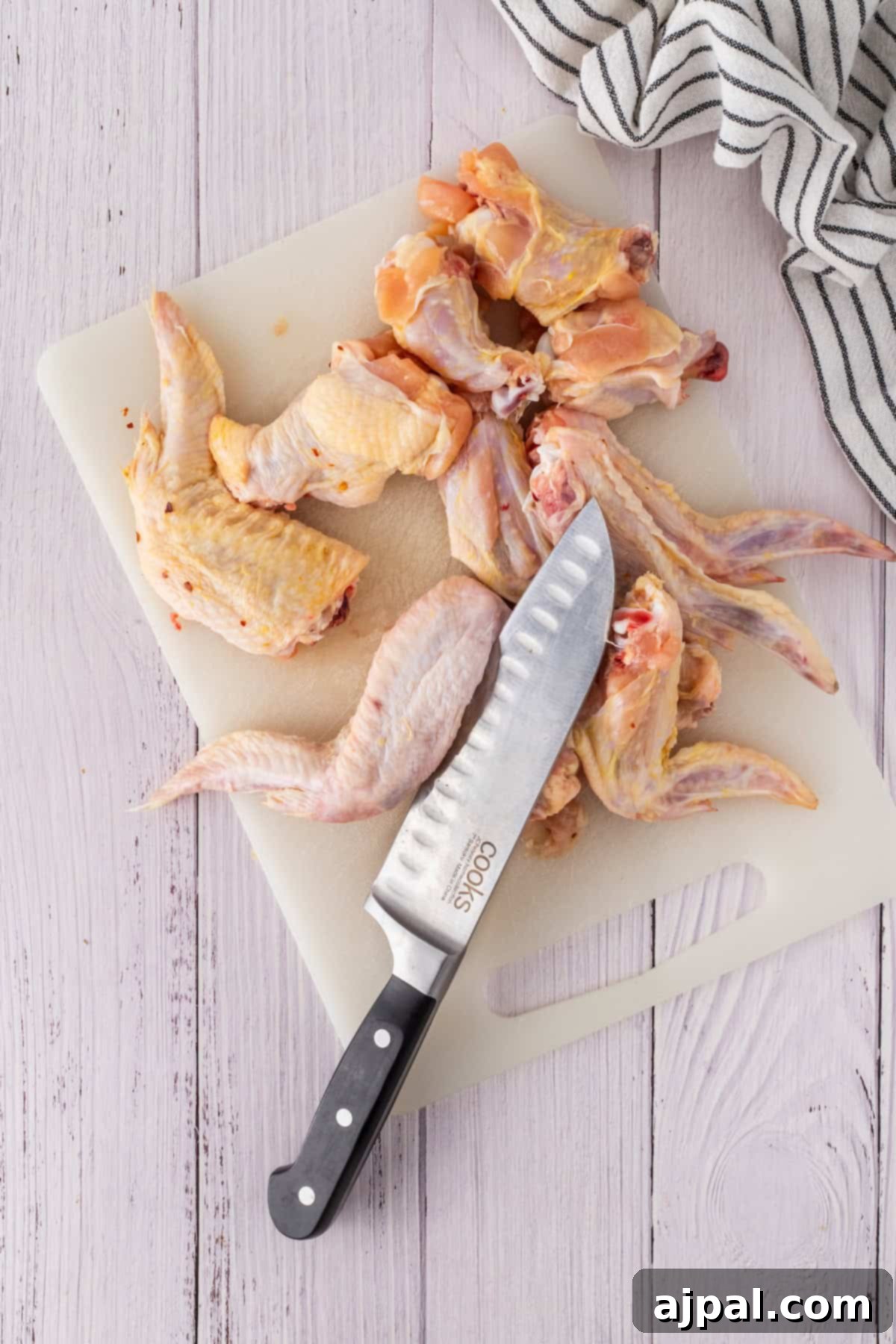 Raw chicken wings and a knife rest on a cutting board, ready for preparation, surrounded by the enticing aroma of garlic on a light wooden surface with a striped cloth nearby.