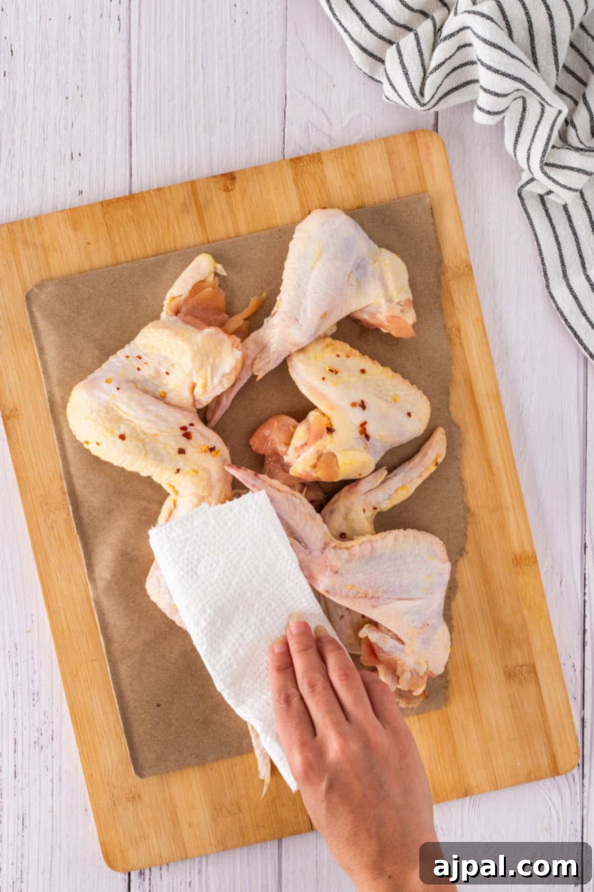 Person patting raw chicken wings dry with a paper towel on a wooden cutting board, preparing them for a delicious honey garlic glaze; white cloth nearby.