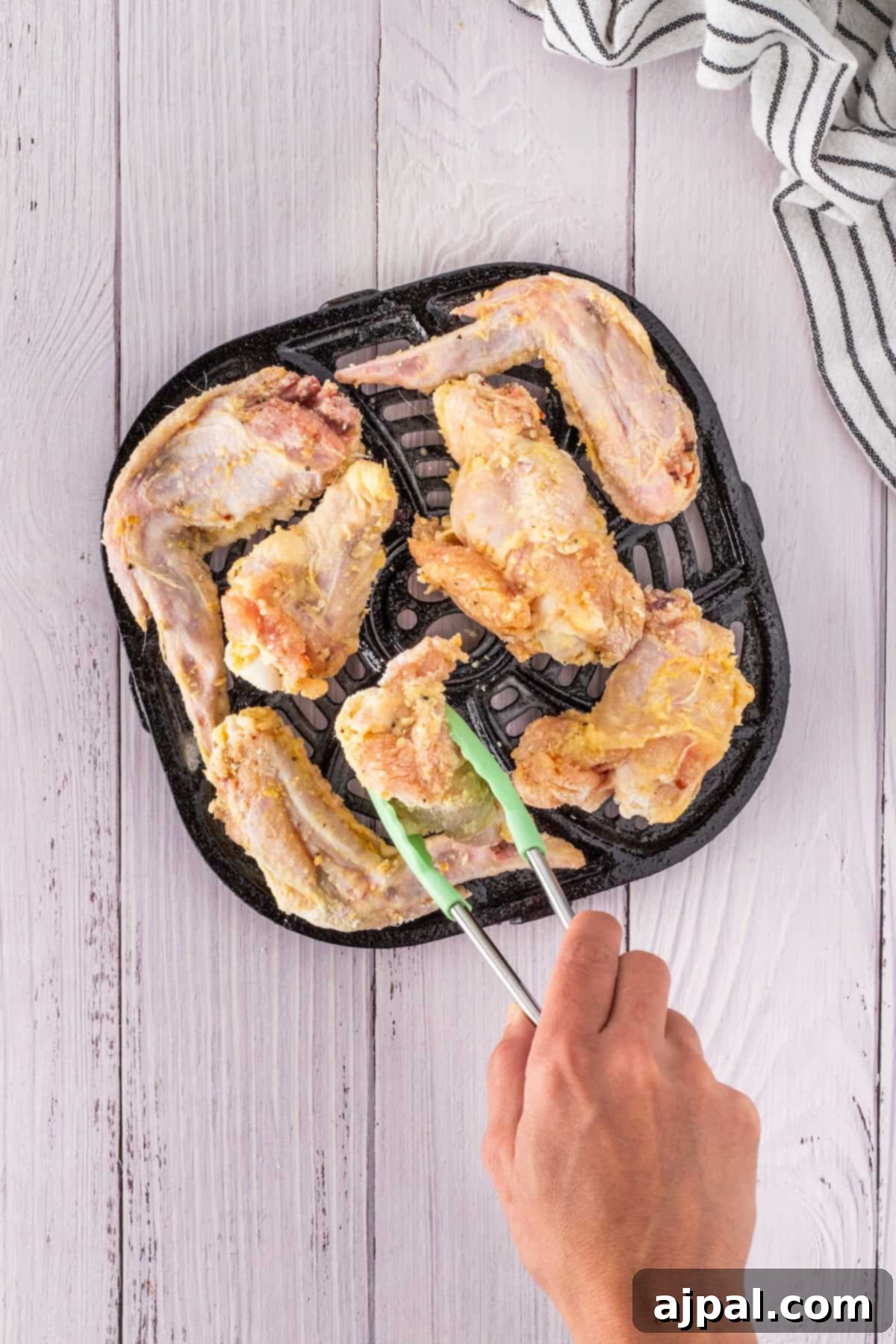 Raw chicken wings on a black grill pan with a hand using tongs to place one piece, set on a light wooden surface with a striped cloth nearby, exude an inviting aroma of garlic.