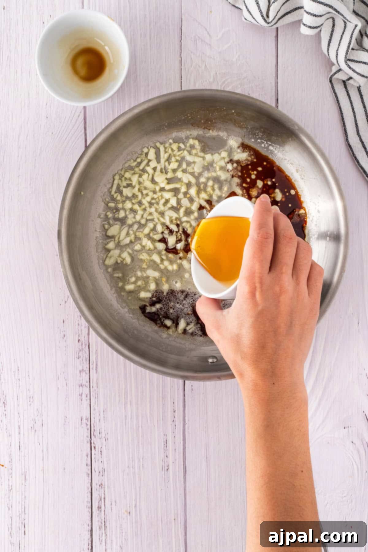 Hand pouring honey from a small white cup into a skillet with chopped garlic and a rich brown liquid. Striped cloth and empty cup sit nearby on the white wooden surface, ready for cooking magic.