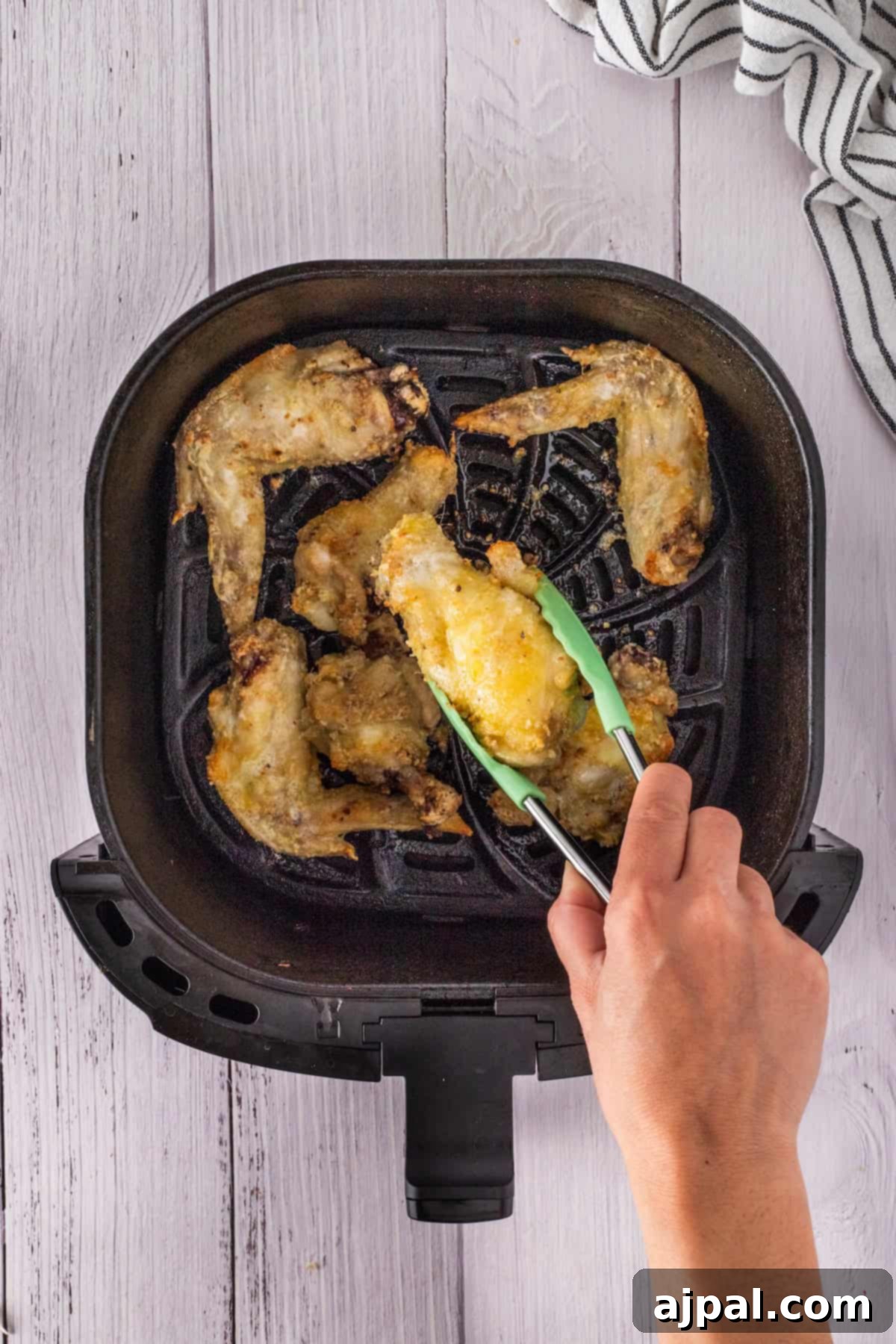 A hand holding green tongs lifts a crispy piece of fried chicken wings from an air fryer filled with delicious pieces. A white and black striped cloth is visible beside the air fryer, hinting at the perfect blend of honey and garlic seasoning.
