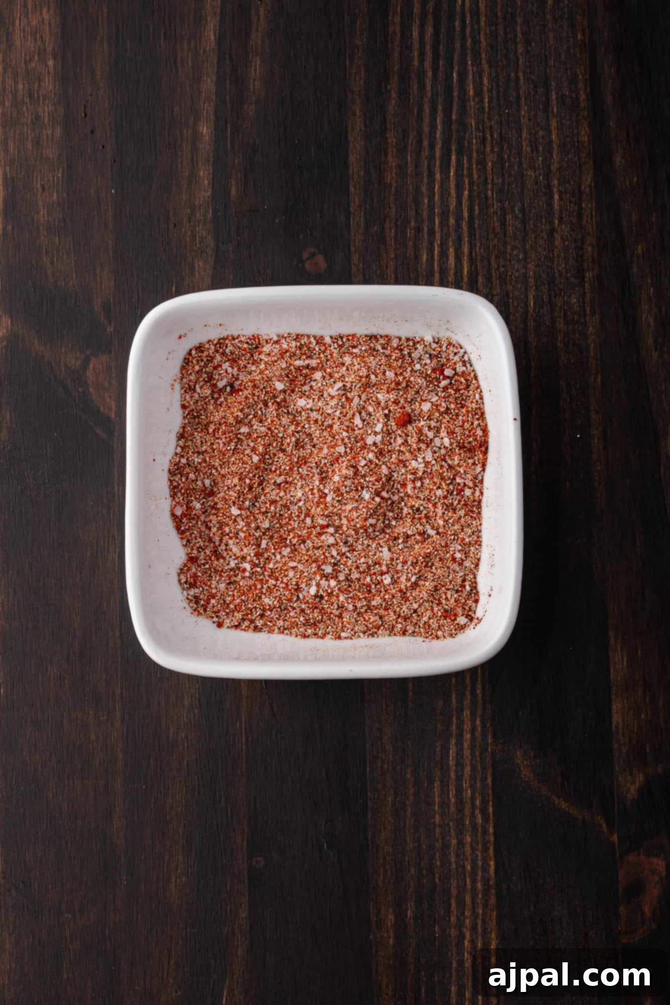 Juicy BBQ Chicken Quarters 6 A white square dish filled with a blend of red and brown spices sits on a dark wooden surface, perfect for seasoning BBQ chicken.