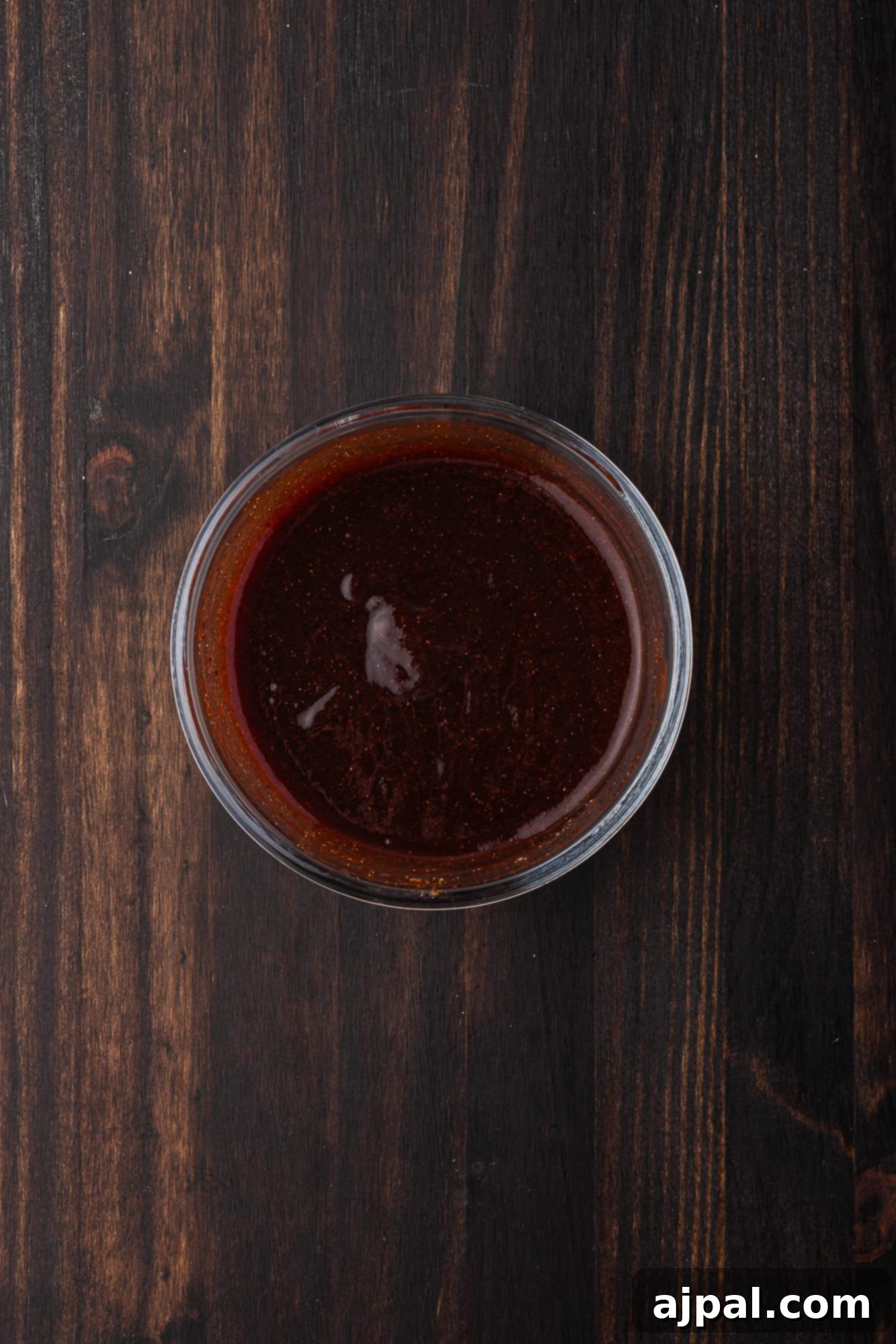 Juicy BBQ Chicken Quarters 9 A small glass bowl filled with rich BBQ sauce rests on a dark wooden surface, perfect for slathering over grilled chicken quarters.