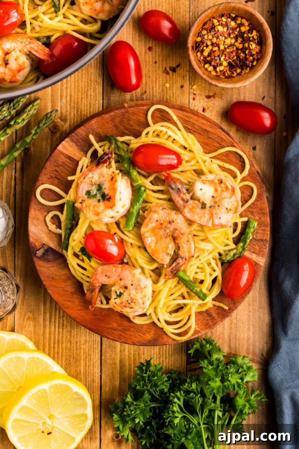 Lemon Garlic Shrimp Pasta 4 Wooden plate with shrimp pasta, cherry tomatoes, and asparagus. Surrounded by lemon slices, parsley, red pepper flakes, fresh garlic cloves, and more cherry tomatoes on a rustic table.