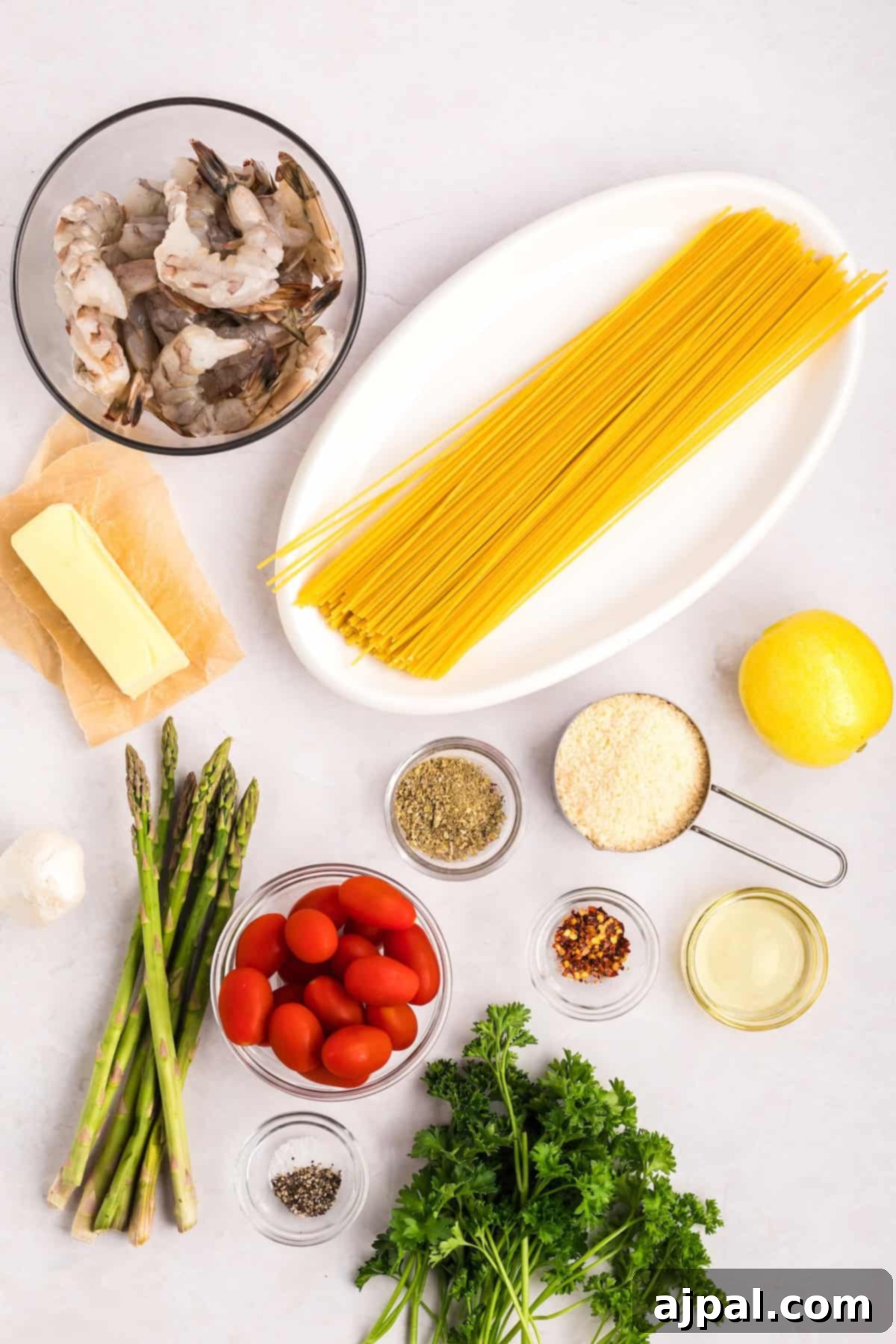 Lemon Garlic Shrimp Pasta 6 Ingredients for a pasta dish are artfully arranged: succulent shrimp, spaghetti, zesty lemon, asparagus, cherry tomatoes, a block of garlic-infused butter, aromatic olive oil, spices, and fresh parsley on a light surface.