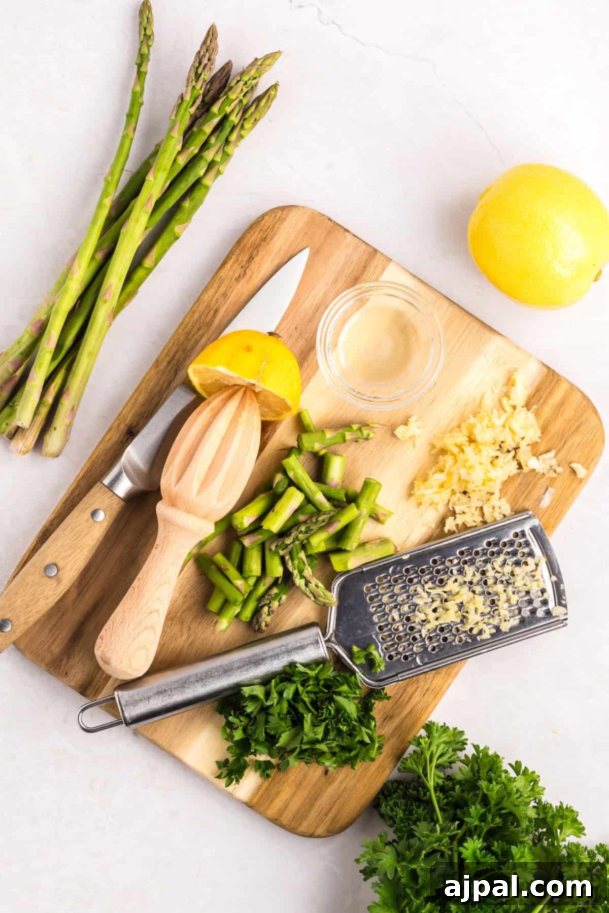 Lemon Garlic Shrimp Pasta 7 Chopped asparagus, garlic, parsley, and a halved lemon rest on a wooden board with a juicer, grater, and knife. A small bowl of liquid sits beside it. Whole asparagus and parsley are nearby, ready to elevate your garlic shrimp pasta into a culinary masterpiece.