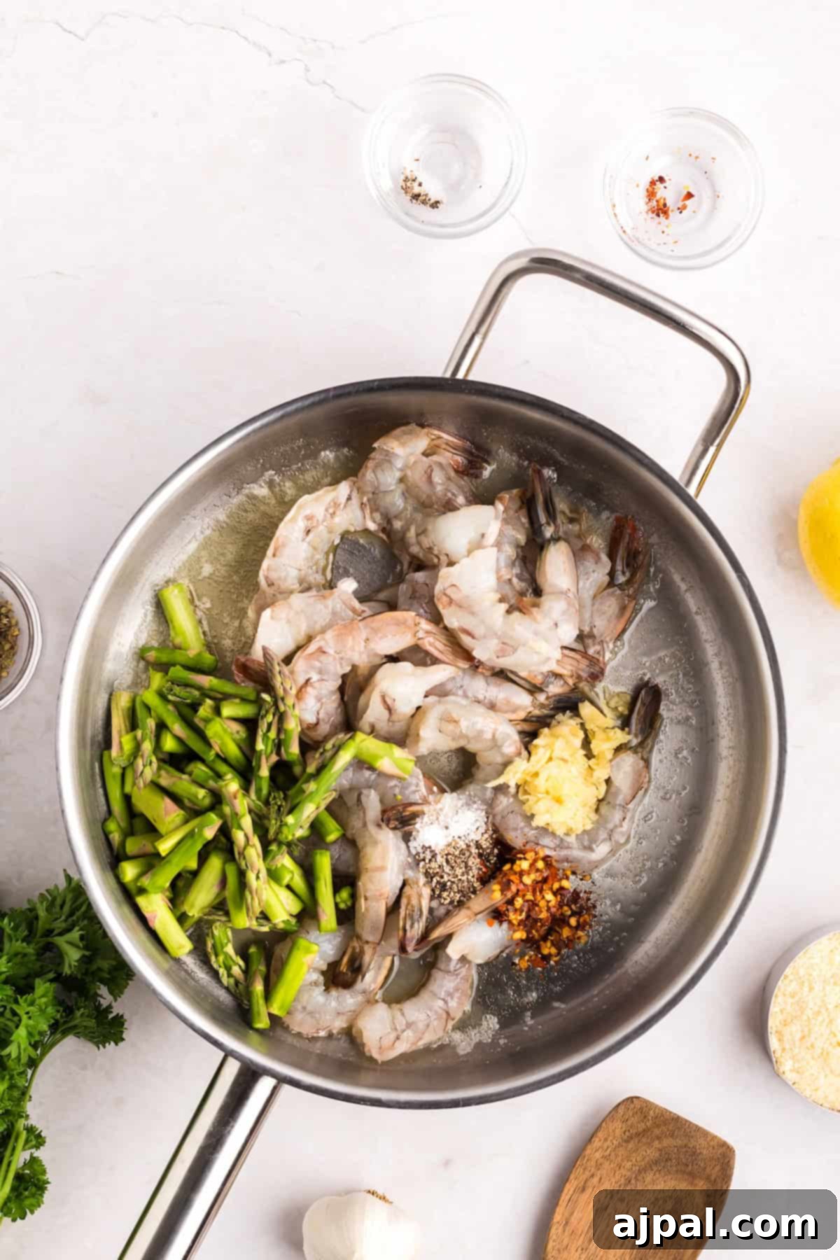 Lemon Garlic Shrimp Pasta 8 Shrimp, asparagus, garlic, and spices sizzle in a stainless steel pan as a medley of parsley and lemon enhance the aroma. Nearby, pasta waits to join the flavorful blend, with a wooden spoon ready to combine these delicate ingredients into a delightful dish.