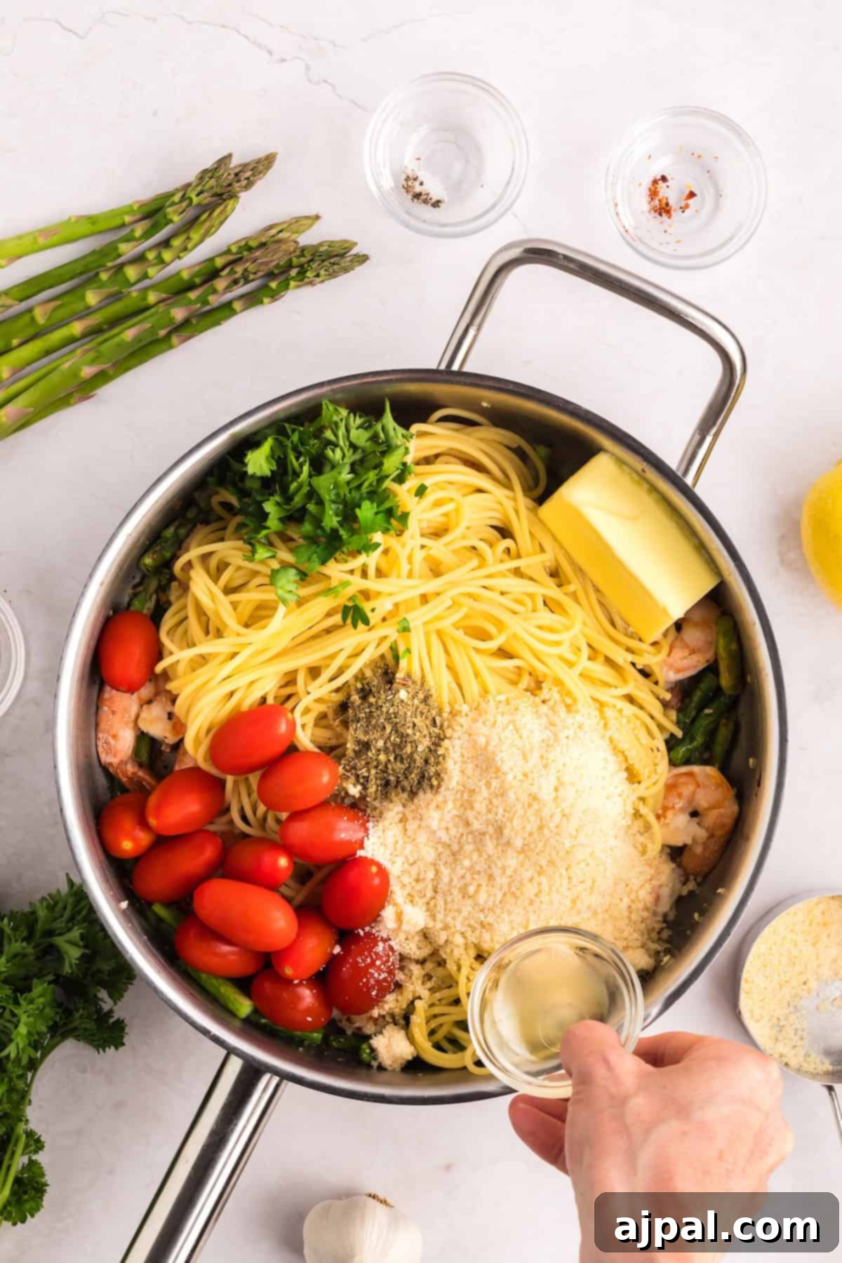 Lemon Garlic Shrimp Pasta 9 A pot with uncooked pasta, garlic-scented shrimp, cherry tomatoes, parsley, asparagus, butter, cheese, and spices. A hand adds liquid from a small glass bowl.