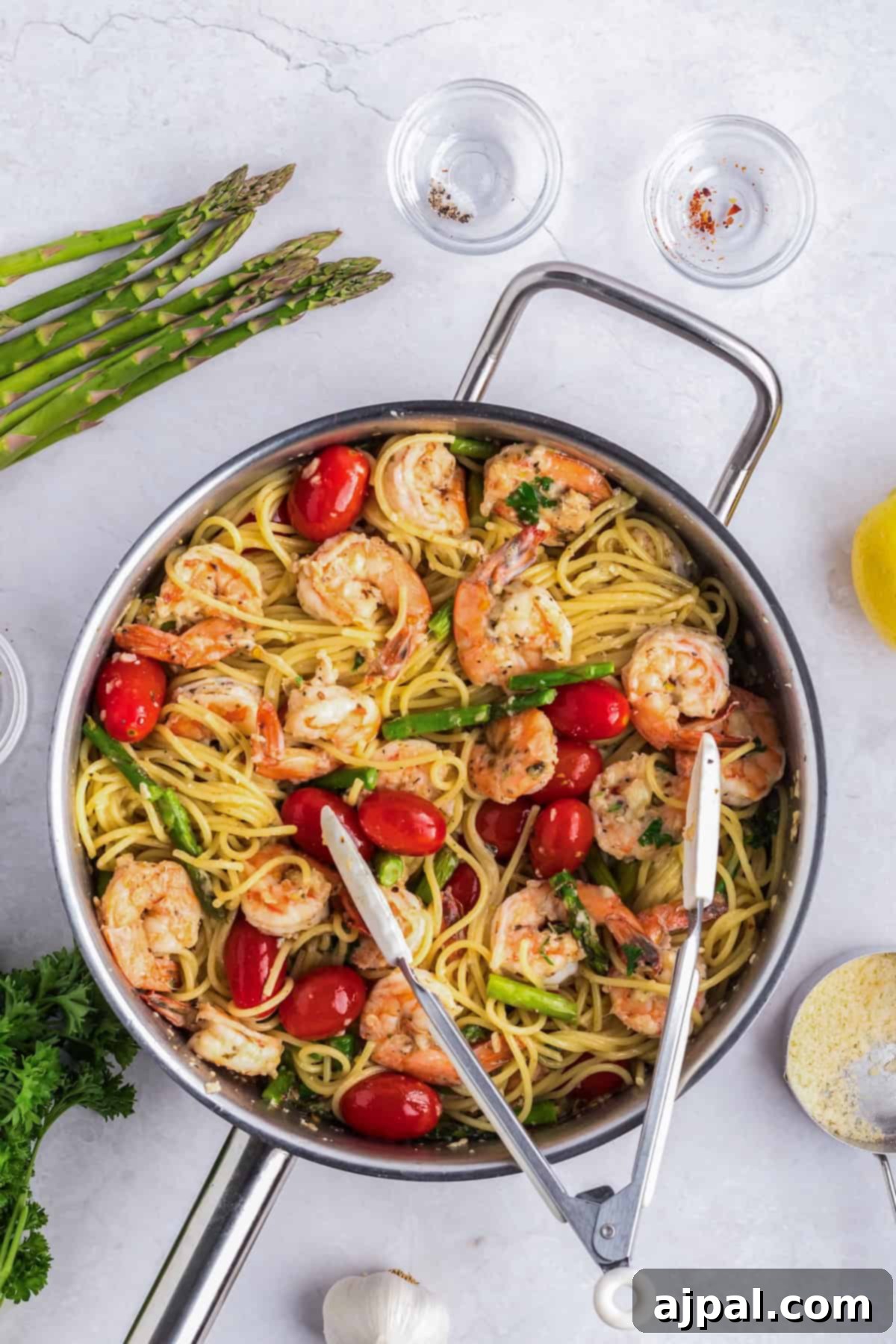 Lemon Garlic Shrimp Pasta 10 A skillet with shrimp scampi pasta showcases succulent shrimp, spaghetti, cherry tomatoes, and asparagus tips. Nearby are parsley, vibrant garlic cloves, lemon wedges, grated cheese, and small bowls of seasonings.