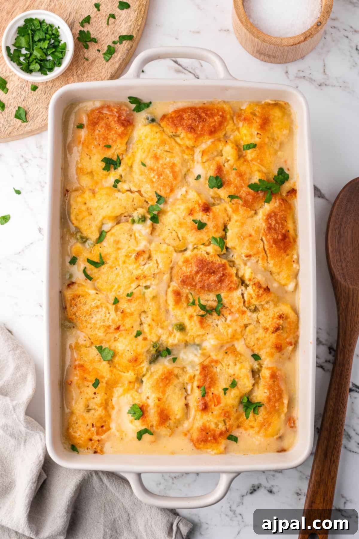Comforting Chicken and Dumpling Bake 11 Golden-brown biscuit-topped chicken casserole nestled in a rectangular dish, garnished with parsley, sits on a marble surface beside a wooden spoon and a small bowl of chopped herbs.
