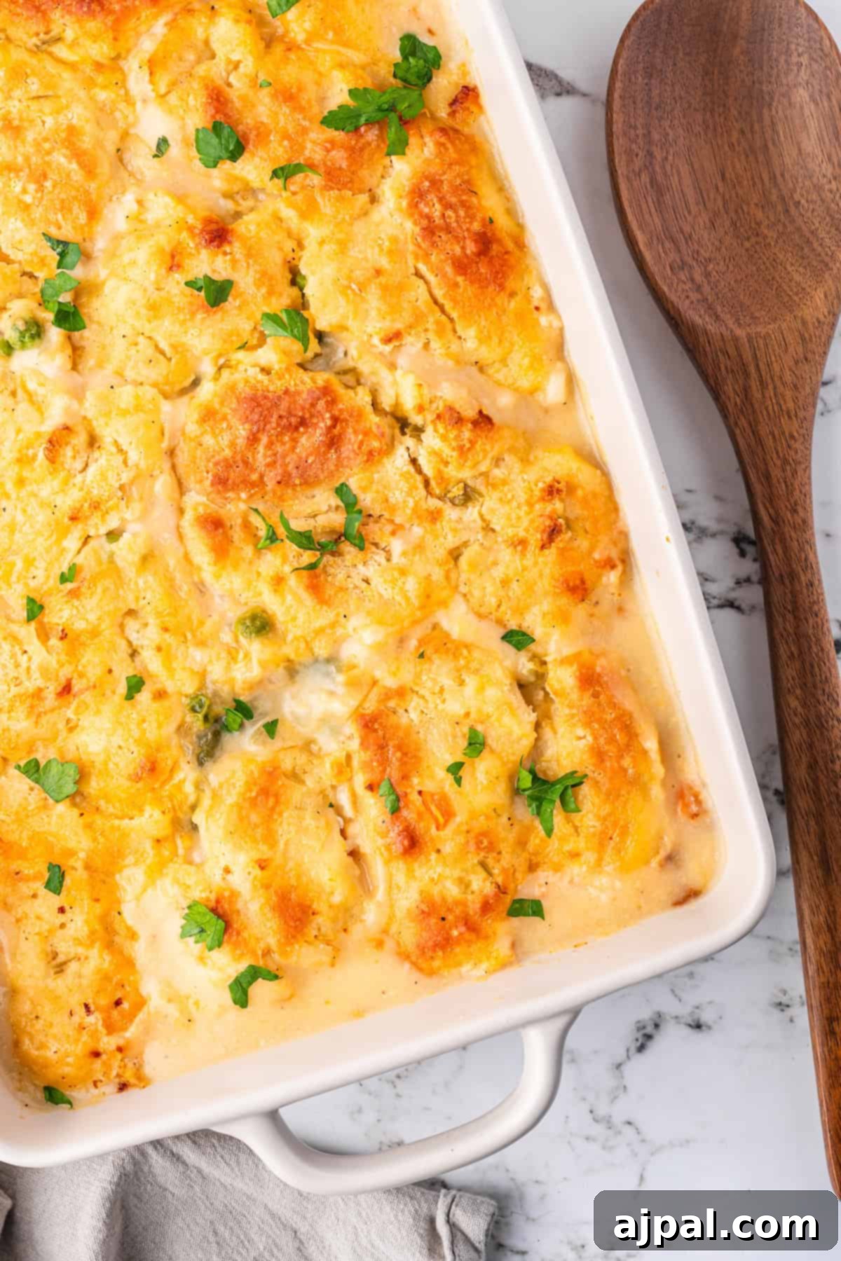 Comforting Chicken and Dumpling Bake 15 Chicken casserole with a golden, cheesy crust and parsley garnish graces a rectangular dish, sitting beside a wooden serving spoon on the marble surface.