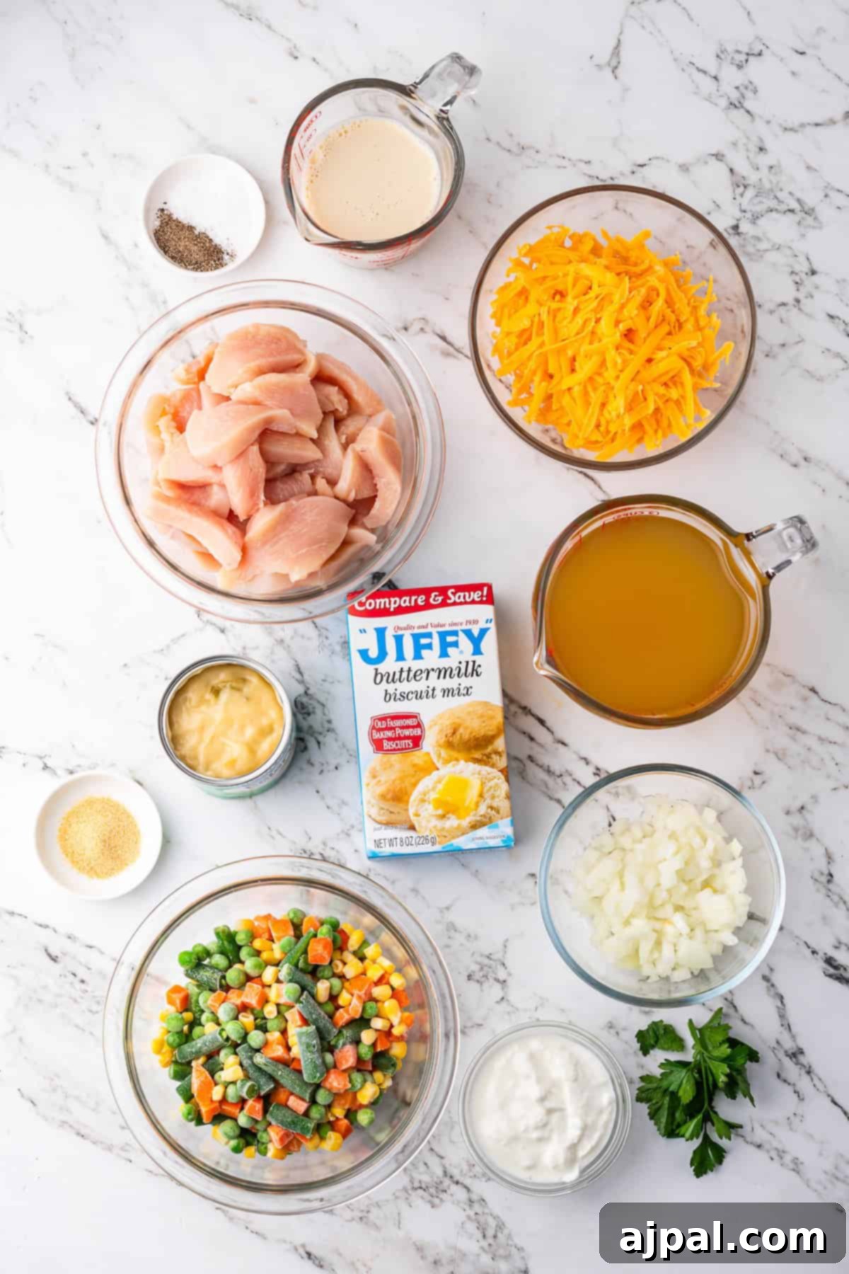Comforting Chicken and Dumpling Bake 3 Ingredients for a chicken pot pie recipe include raw chicken, shredded cheese, broth, cream, diced onions, mixed vegetables, buttermilk biscuit mix, sour cream, pepper, and parsley on a marble surface.