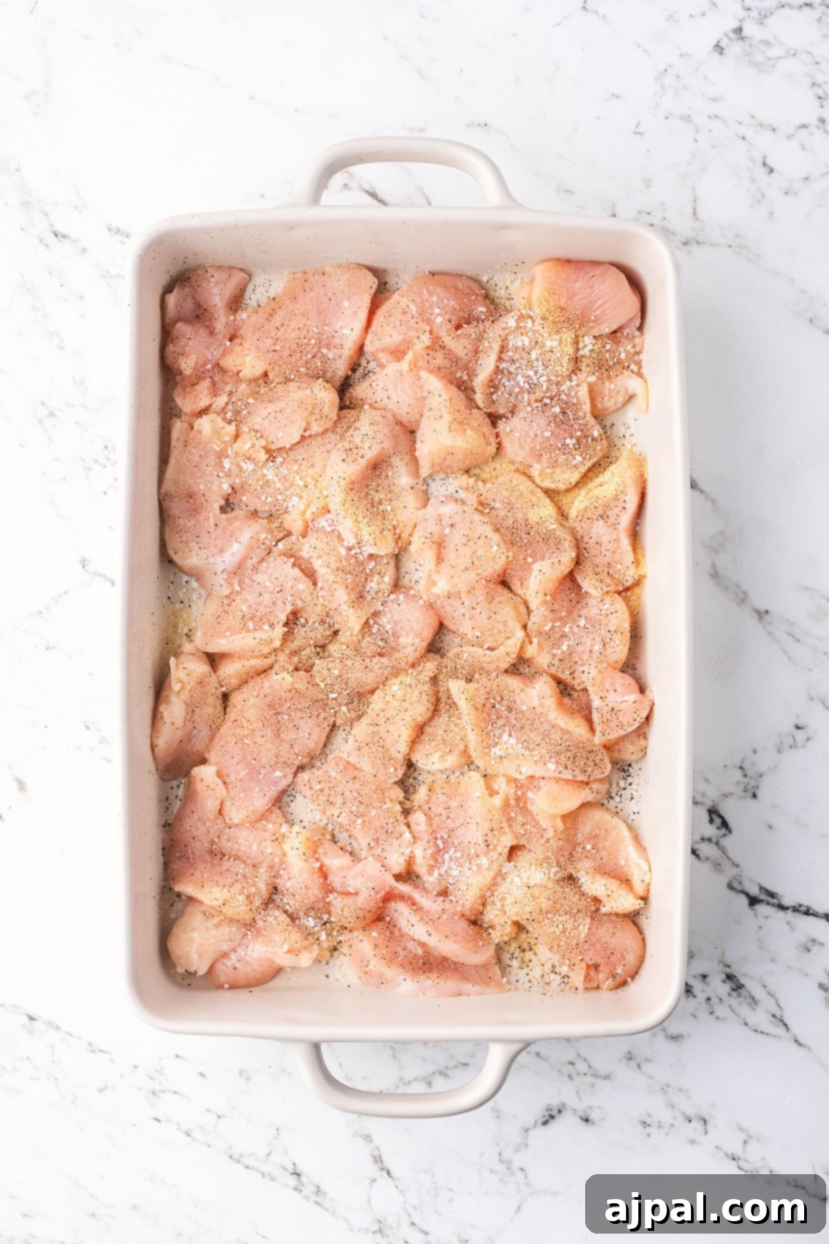 Comforting Chicken and Dumpling Bake 4 A rectangular white baking dish filled with raw chicken pieces, seasoned with spices, sits on a marble countertop, ready to transform into a delicious casserole.