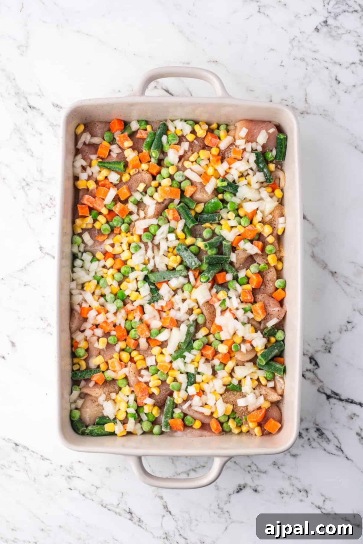 Comforting Chicken and Dumpling Bake 5 Mixed vegetables and diced onions are spread over a layer of chicken in a baking dish, preparing for the next steps of a flavorful casserole.