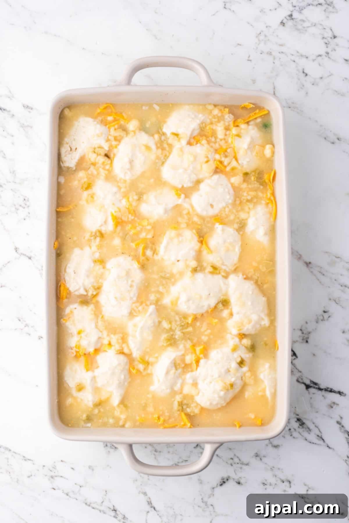 Comforting Chicken and Dumpling Bake 10 A baking dish is filled with a creamy mixture, reminiscent of a comforting chicken pot pie, and topped with white, fluffy dollops resembling dumplings. It’s garnished with corn and small green pieces, all set elegantly on a marble surface.