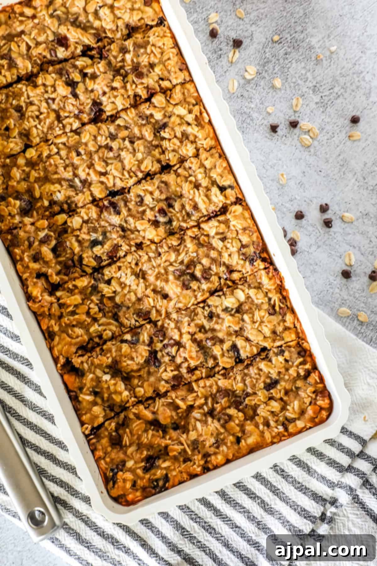 Artisan Sourdough Granola Bites 2 A tray of sourdough granola bars with visible chocolate chips, neatly cut into rectangles, sits on a striped kitchen towel with some oats scattered nearby.