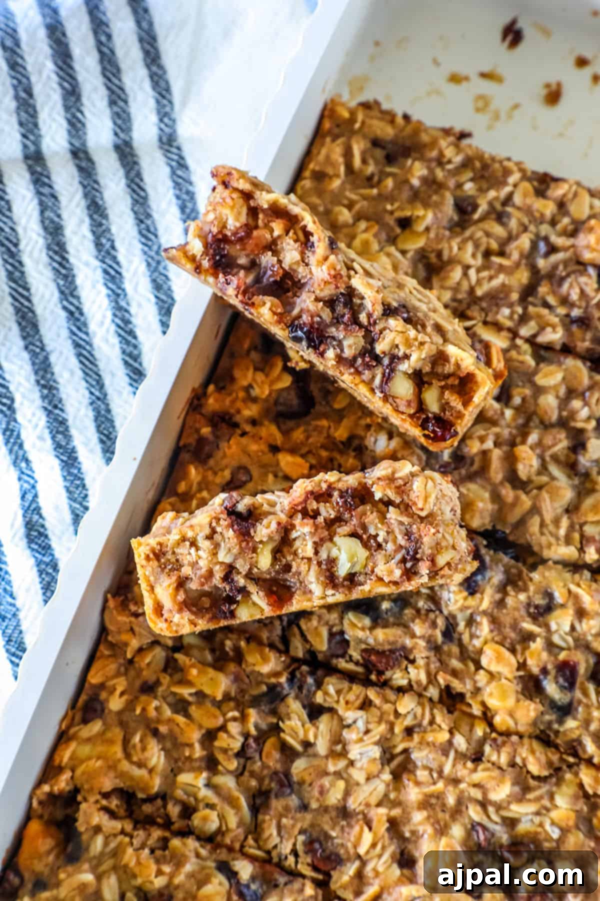 Artisan Sourdough Granola Bites 3 A close-up of homemade sourdough granola bars with visible nuts and dried fruit, cut into pieces in a white baking dish next to a striped kitchen towel.