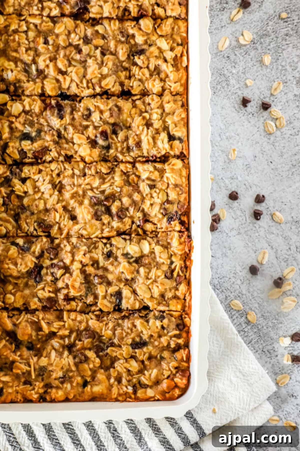 Artisan Sourdough Granola Bites 4 A rectangular baking dish filled with sliced sourdough granola bars sits on a gray textured surface with scattered oats and chocolate chips nearby.