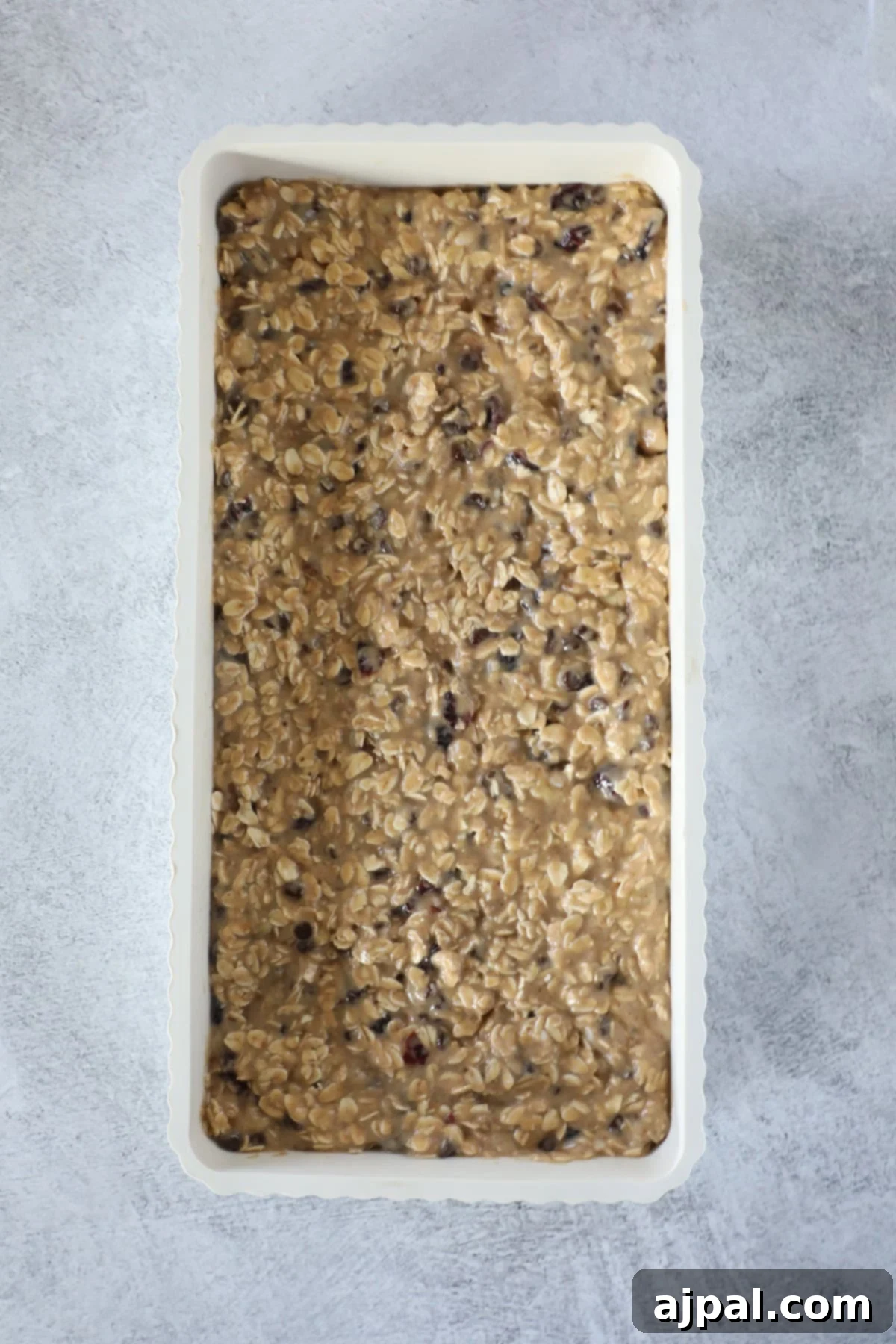 Artisan Sourdough Granola Bites 6 A rectangular pan filled with unbaked sourdough granola bar batter, blending oatmeal and raisins, sits on a gray countertop.