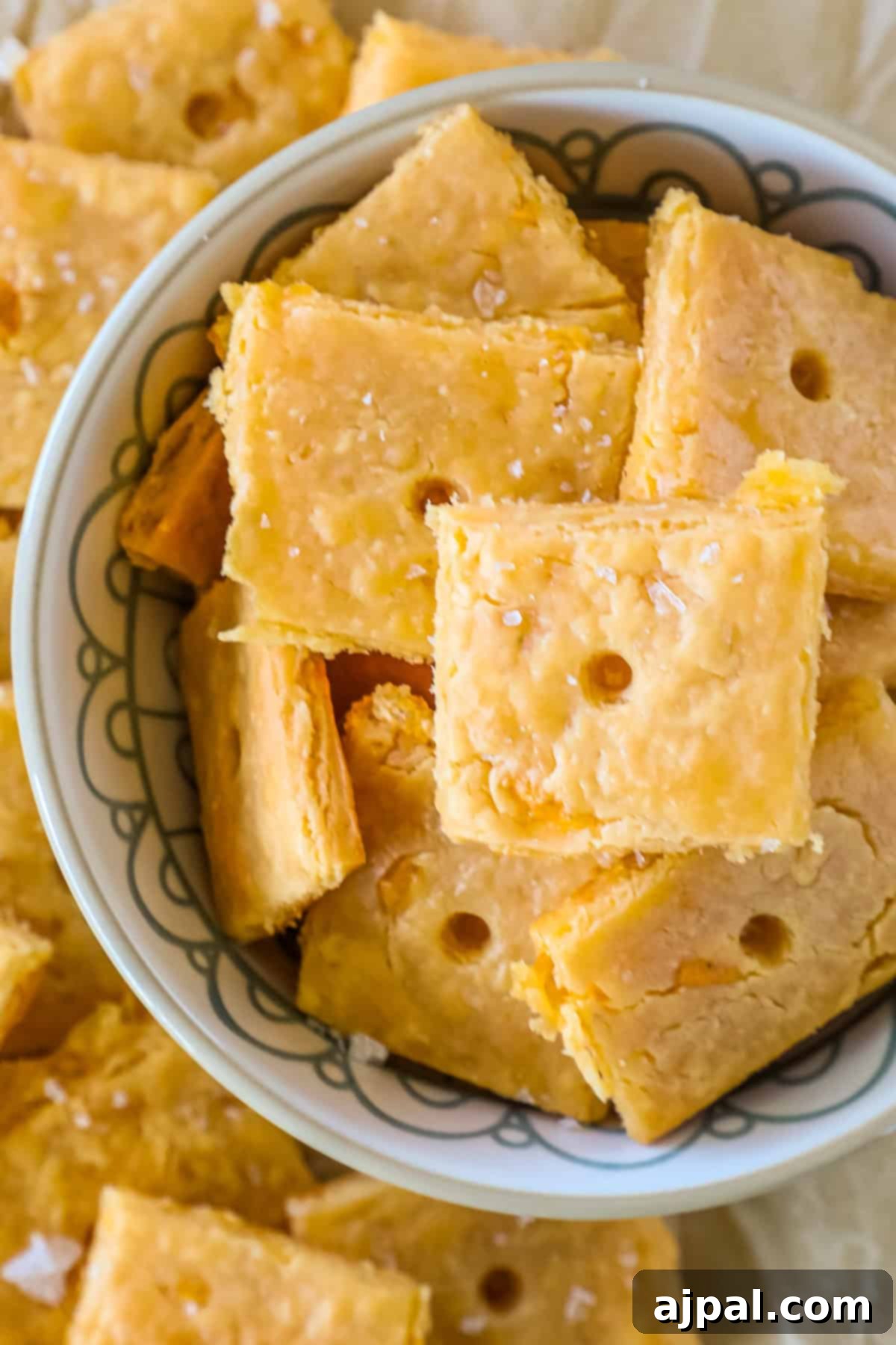 Cheesy Sourdough Discard Crisps 3 A bowl filled with square, golden crackers with visible sea salt flakes on top, surrounded by more crackers.