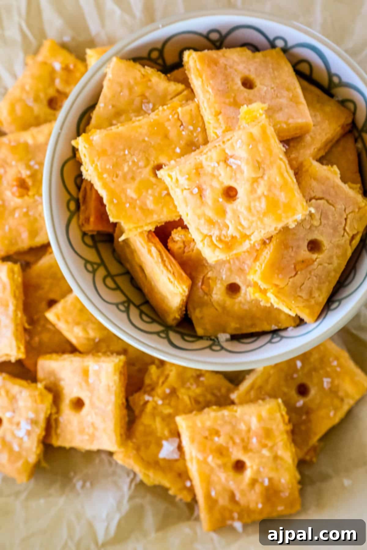 Cheesy Sourdough Discard Crisps 8 A bowl of square, orange-colored cheese crackers crafted with sourdough discard sits on a sheet of parchment paper, surrounded by more scattered crackers.