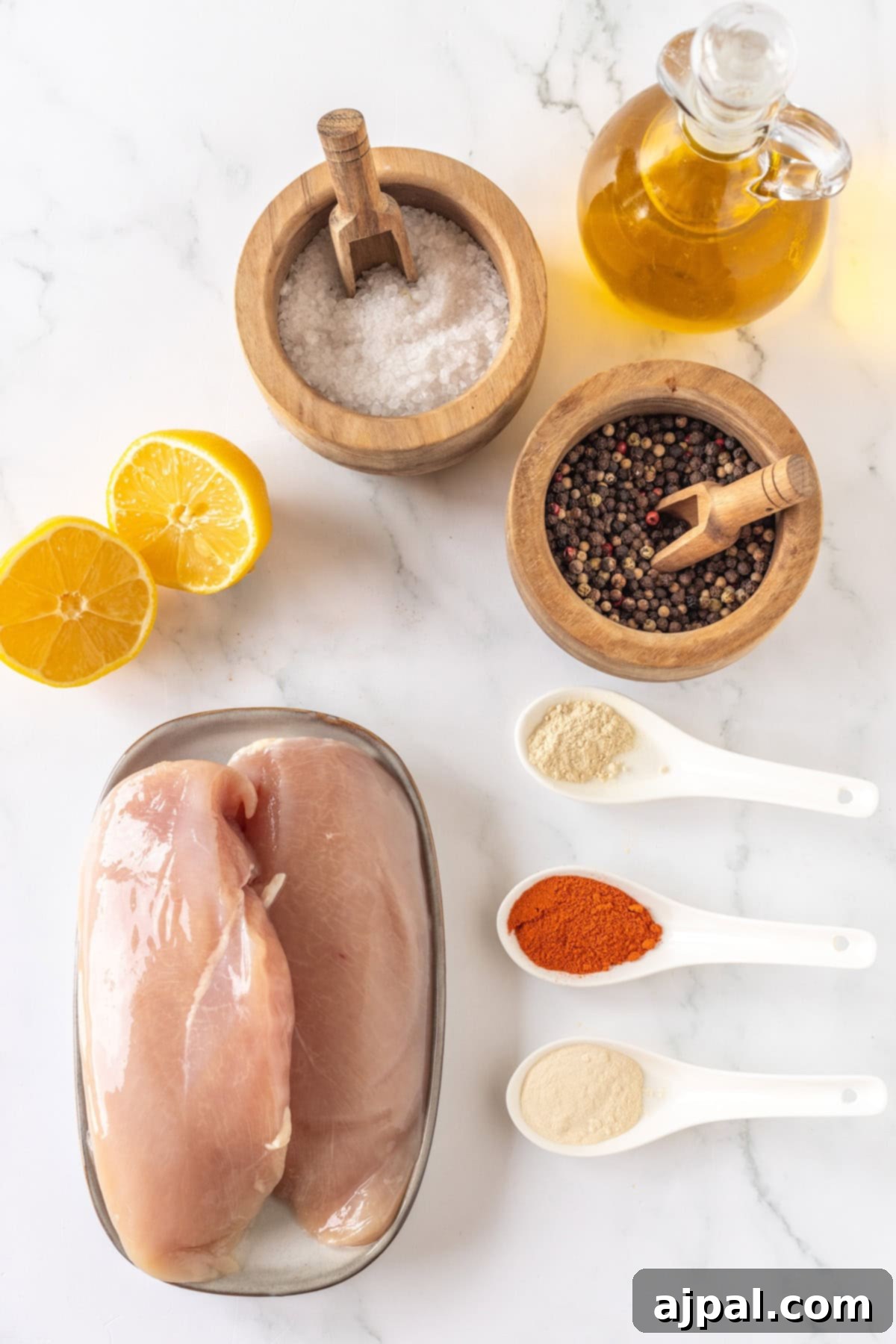 Raw chicken breasts, lemon halves, and olive oil rest in a wooden bowl, accompanied by a sprinkle of salt and peppercorns. Three white spoons hold an array of spices on the marble surface, ready to transform these ingredients into a delectable thin baked chicken masterpiece.