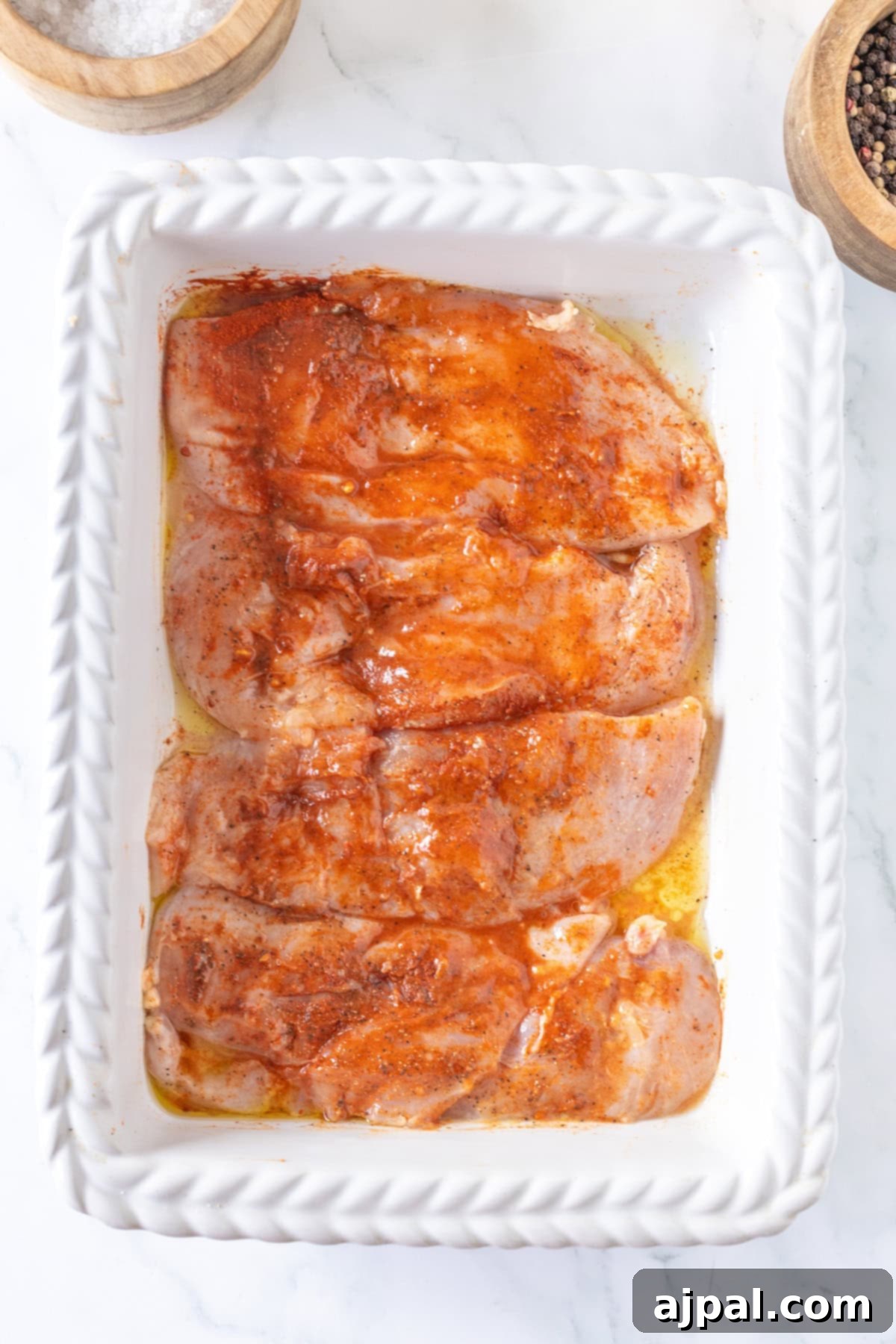 Thin chicken breasts are marinated with spices and oil in a white baking dish, ready to transform into one of your go-to chicken recipes, perfectly set on a marble surface.