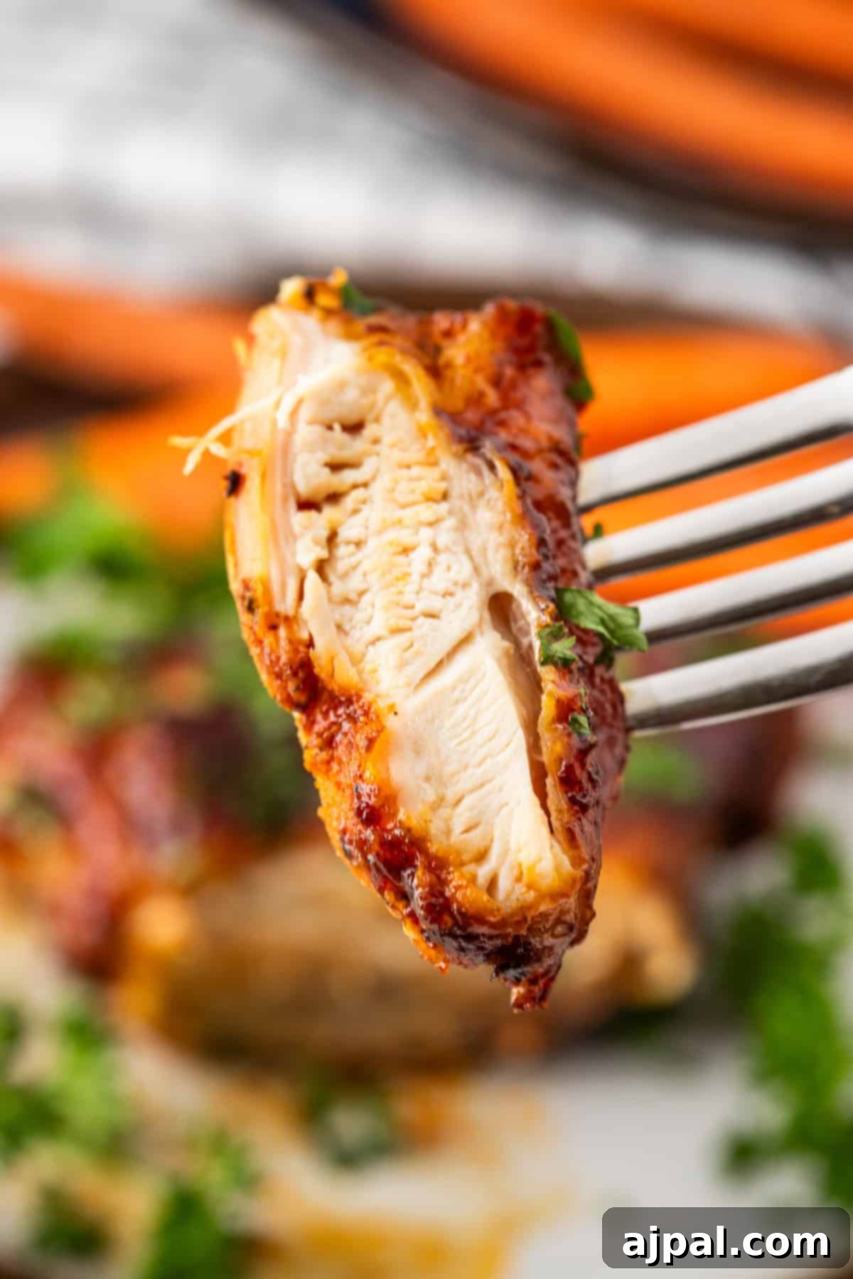 Crispy Air Fryer BBQ Chicken Thighs 2 Close-up of a fork holding a piece of seasoned, cooked BBQ chicken with visible grill marks, against a blurred background of more succulent chicken thighs and green garnish.