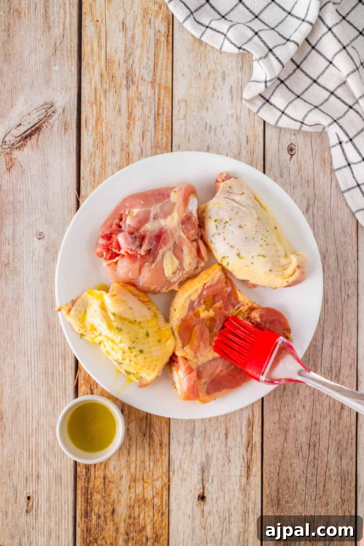 Crispy Air Fryer BBQ Chicken Thighs 4 Raw chicken thighs on a white plate are being brushed with tangy BBQ marinade. A small bowl of marinade sits nearby. The plate rests on a wooden surface with a checkered cloth beside it, ready for the air fryer magic.