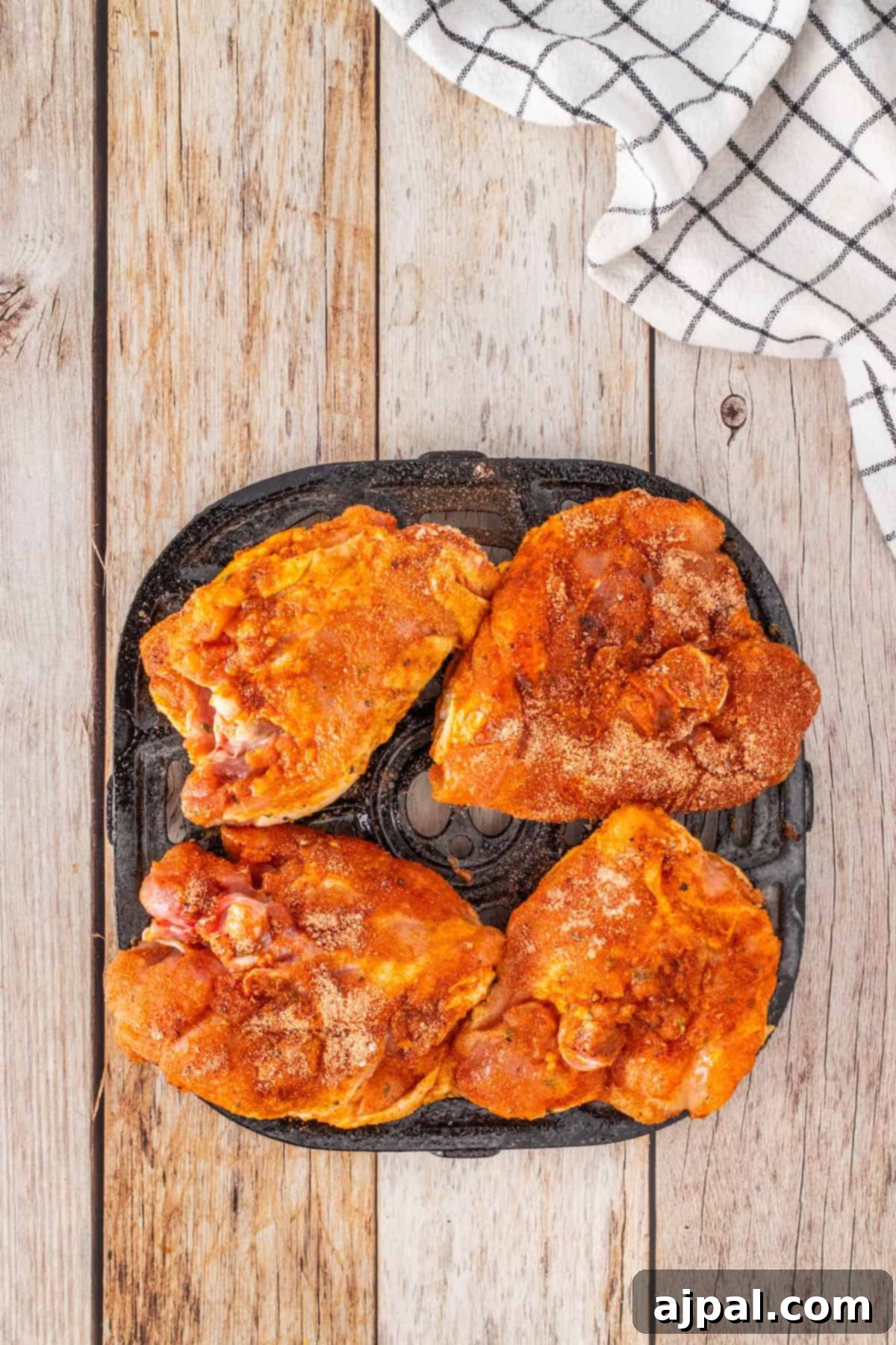 Crispy Air Fryer BBQ Chicken Thighs 6 Four seasoned chicken thighs rest on a black grill pan, ready to become delicious BBQ chicken, placed on a wooden surface with a checkered cloth nearby.