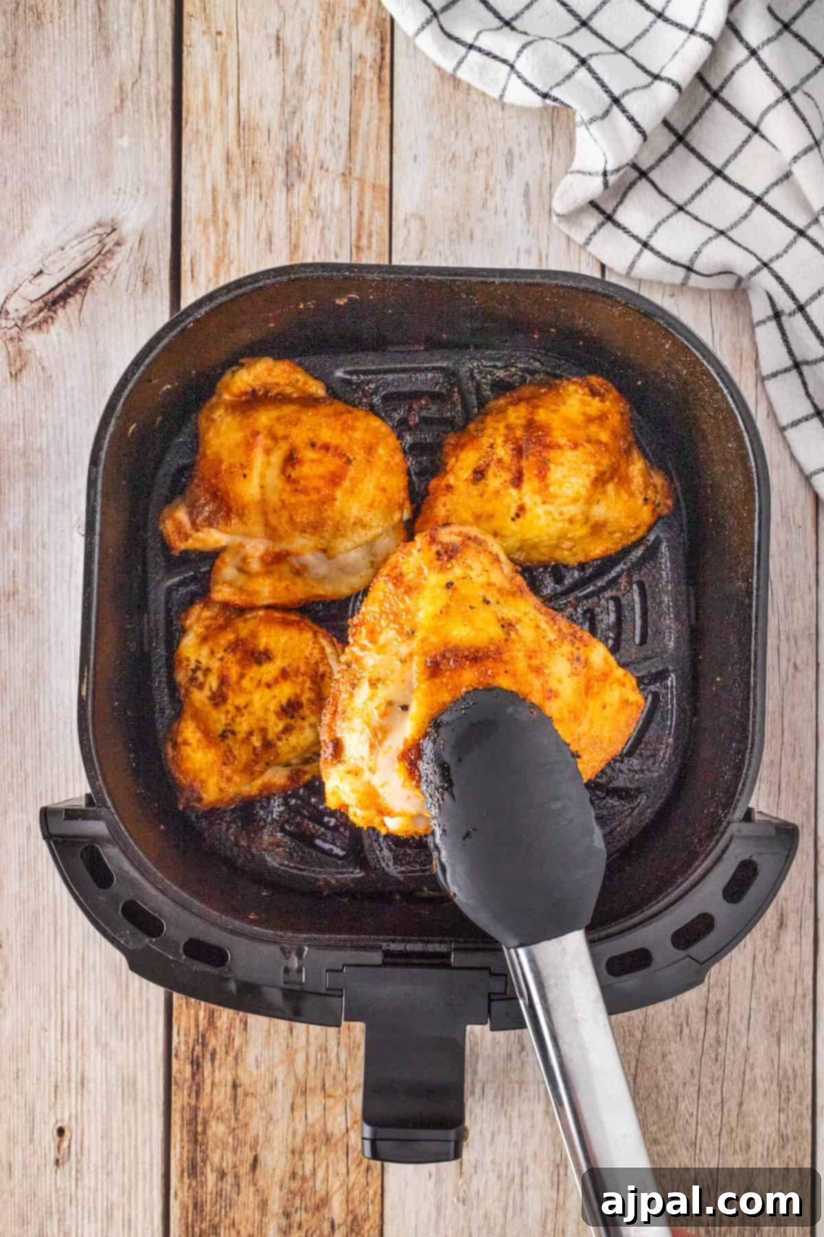 Crispy Air Fryer BBQ Chicken Thighs 7 Four tender BBQ chicken thighs rest in an air fryer basket, with tongs holding one piece, against a wooden surface next to a checkered cloth.