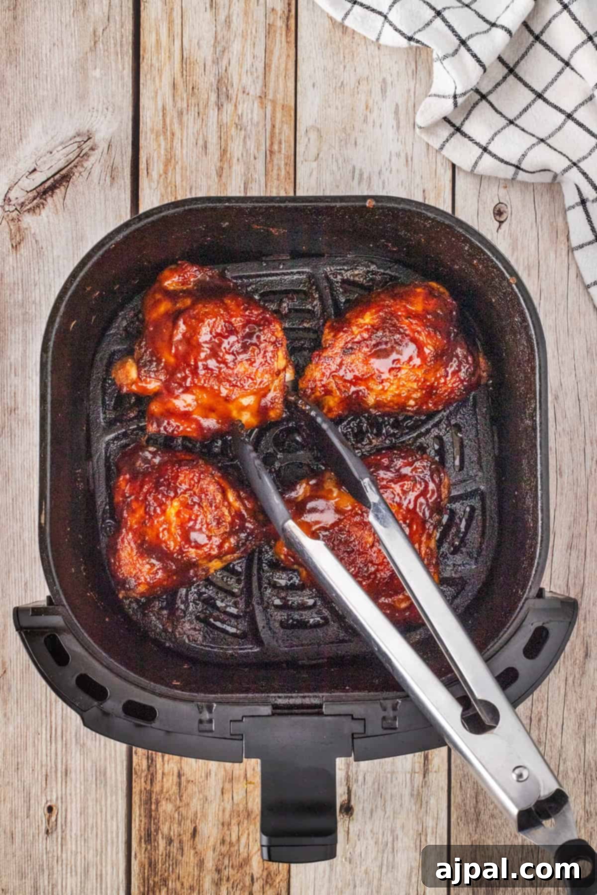 Crispy Air Fryer BBQ Chicken Thighs 8 Three juicy pieces of glazed BBQ chicken thighs sit in an air fryer basket, with stainless steel tongs grasping one piece. A checkered towel is partially visible on the wooden surface beside the fryer.
