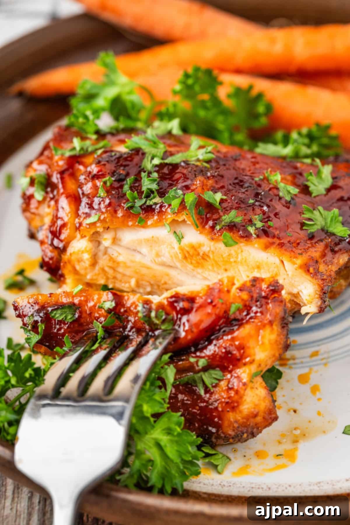 Crispy Air Fryer BBQ Chicken Thighs 10 Close-up of glazed roast chicken thighs on a plate, garnished with parsley and perfectly pierced by a fork. In the background, fresh carrots add color to the scene.